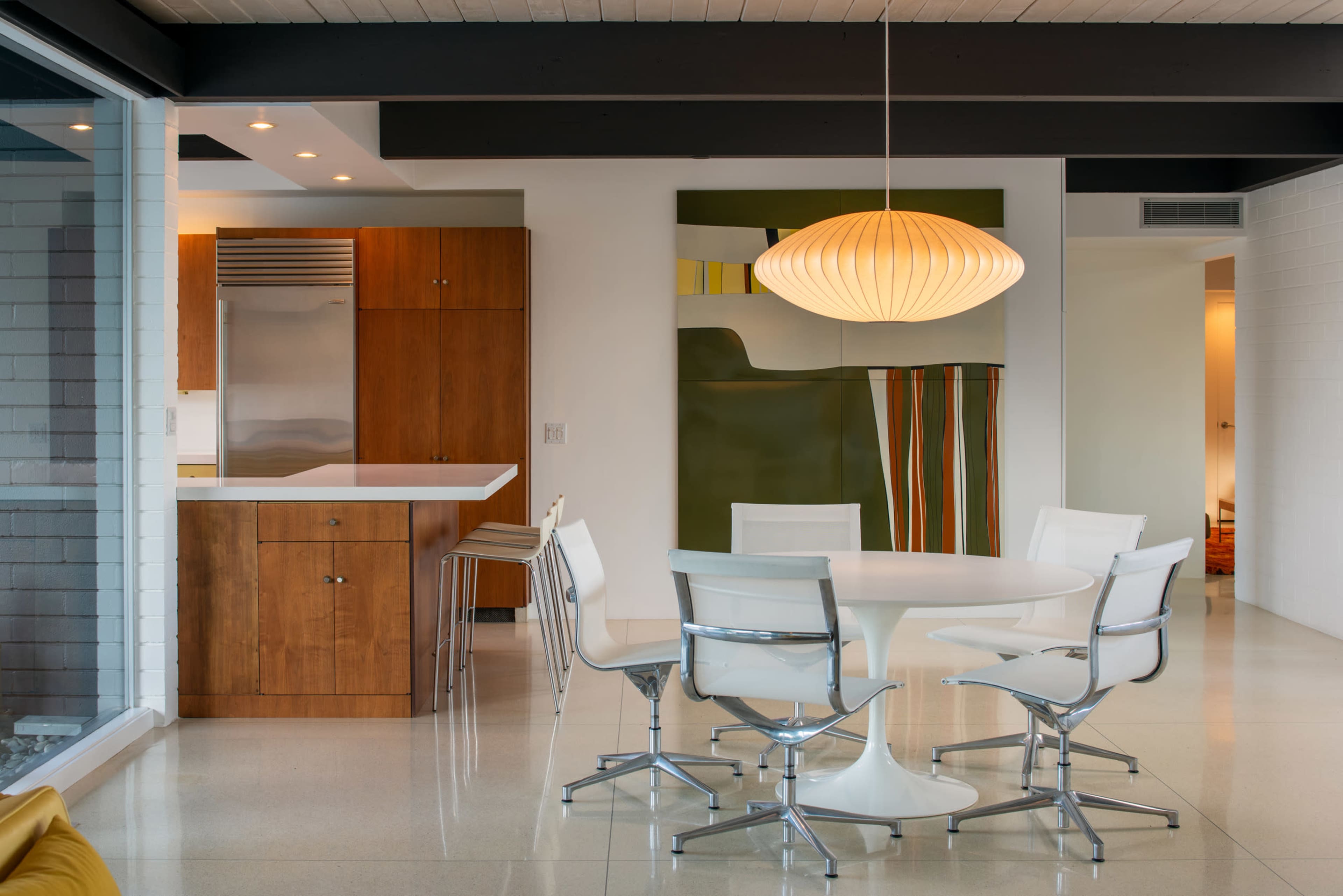 A modern dining area features a round white table surrounded by six sleek chairs, with a wooden kitchen cabinetry backdrop and an abstract wall painting.