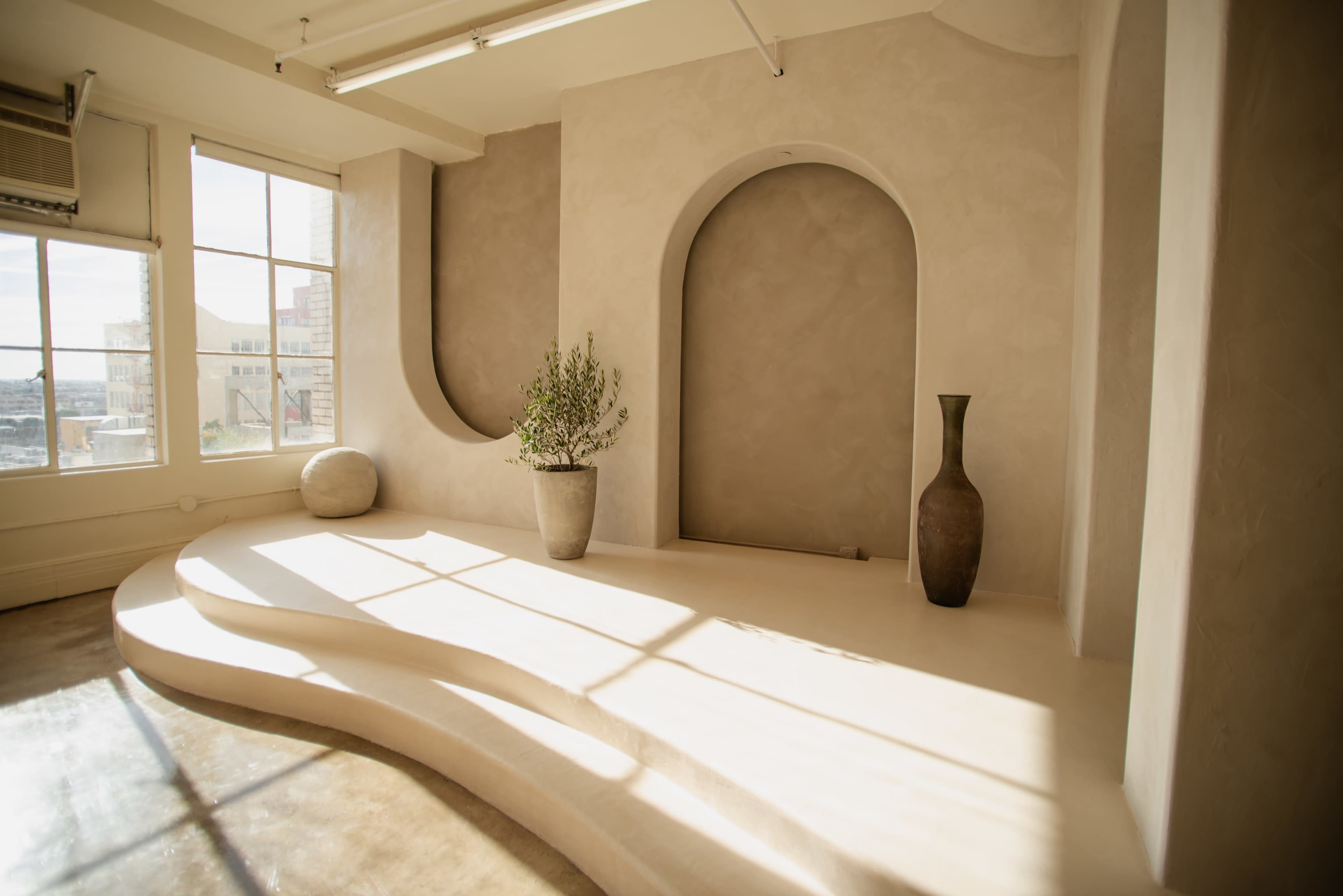 A minimalist interior space with curved walls, a large window, and a vase containing greenery on a sculptural, tiered platform.