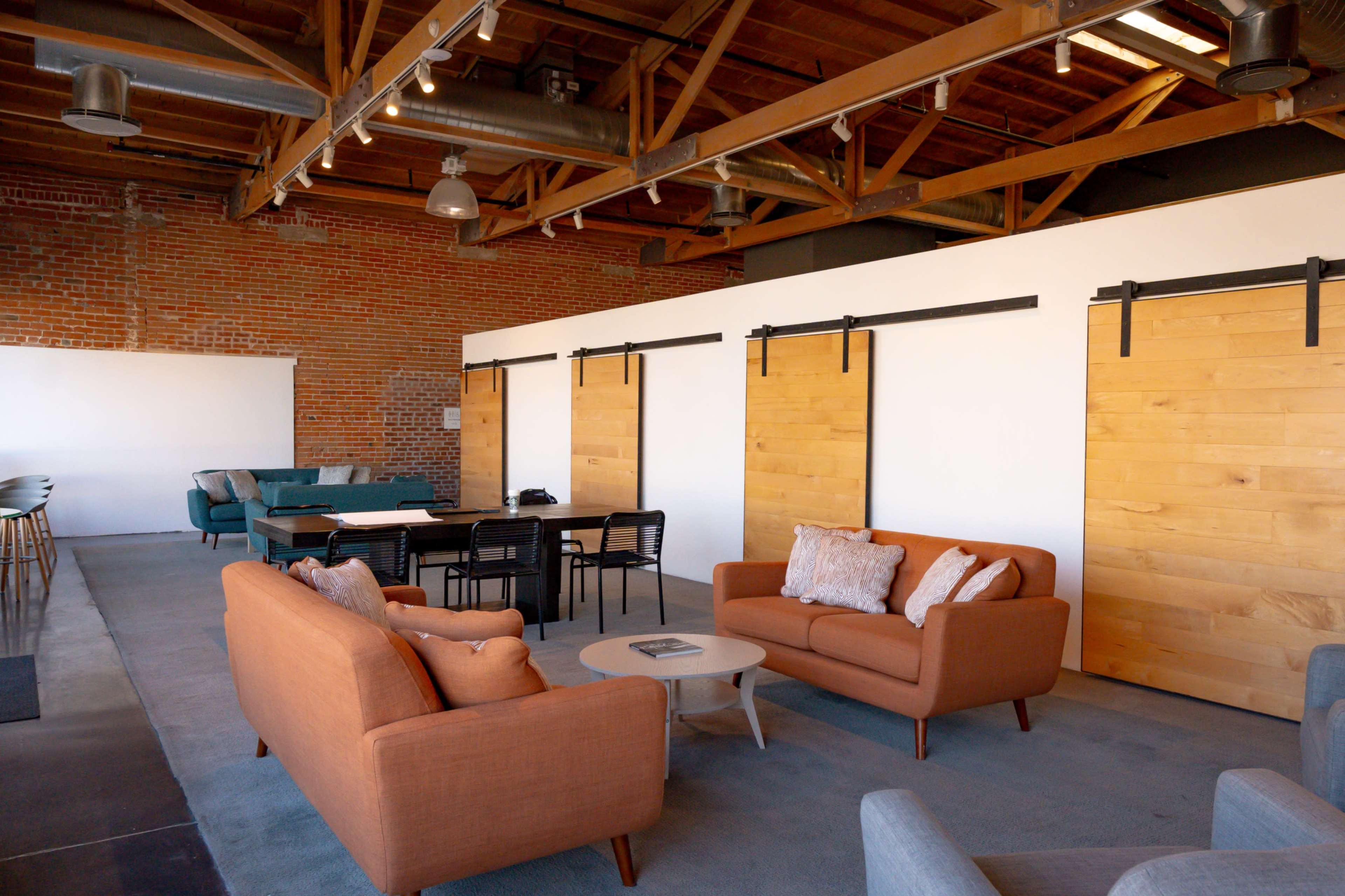 A modern, open-concept office space featuring a seating area with orange and gray sofas, a central table with black chairs, and sliding wooden barn doors against a brick wall.