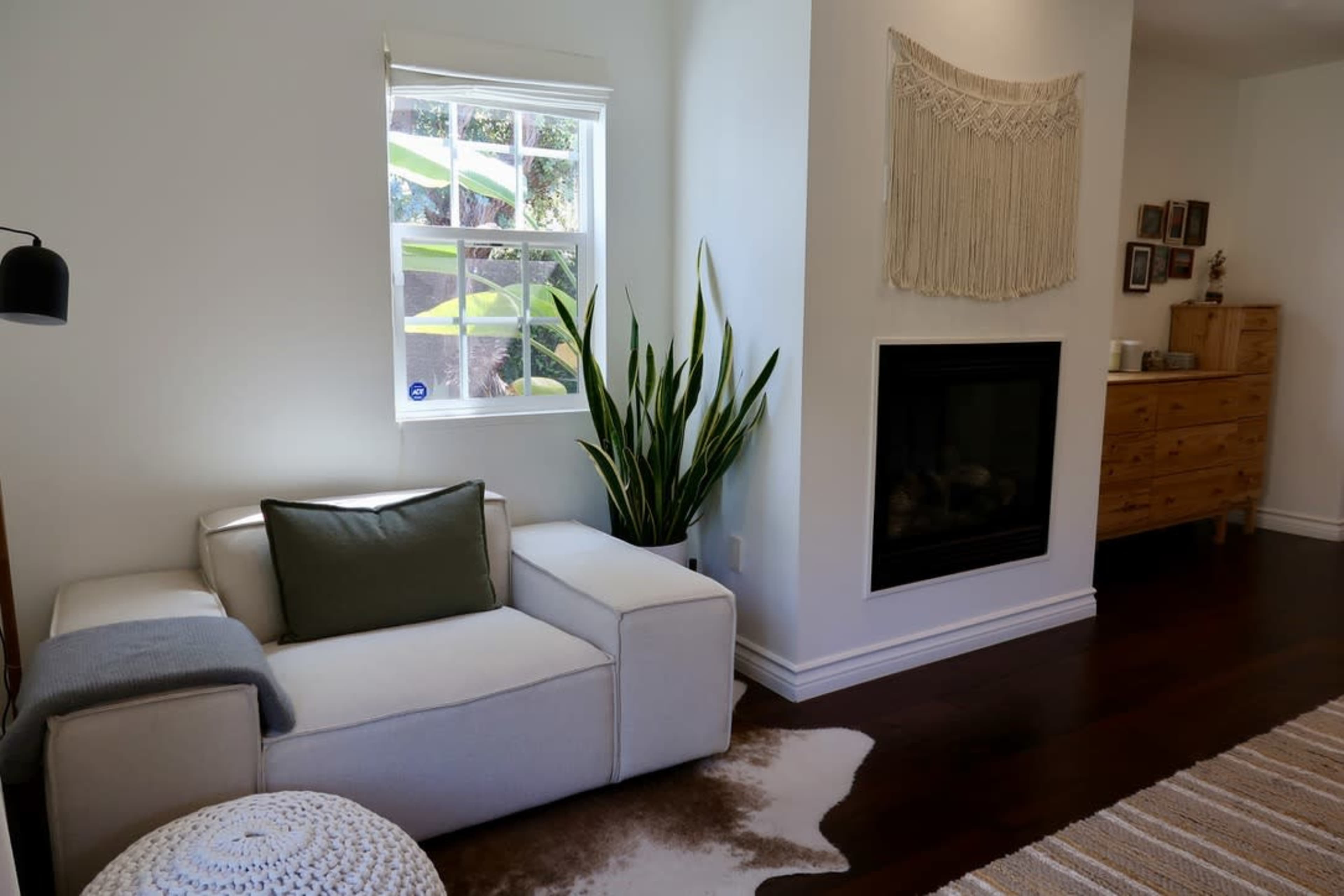 The image shows a modern living room with a white sectional sofa, a potted plant, a fireplace, and a decorative wall hanging.