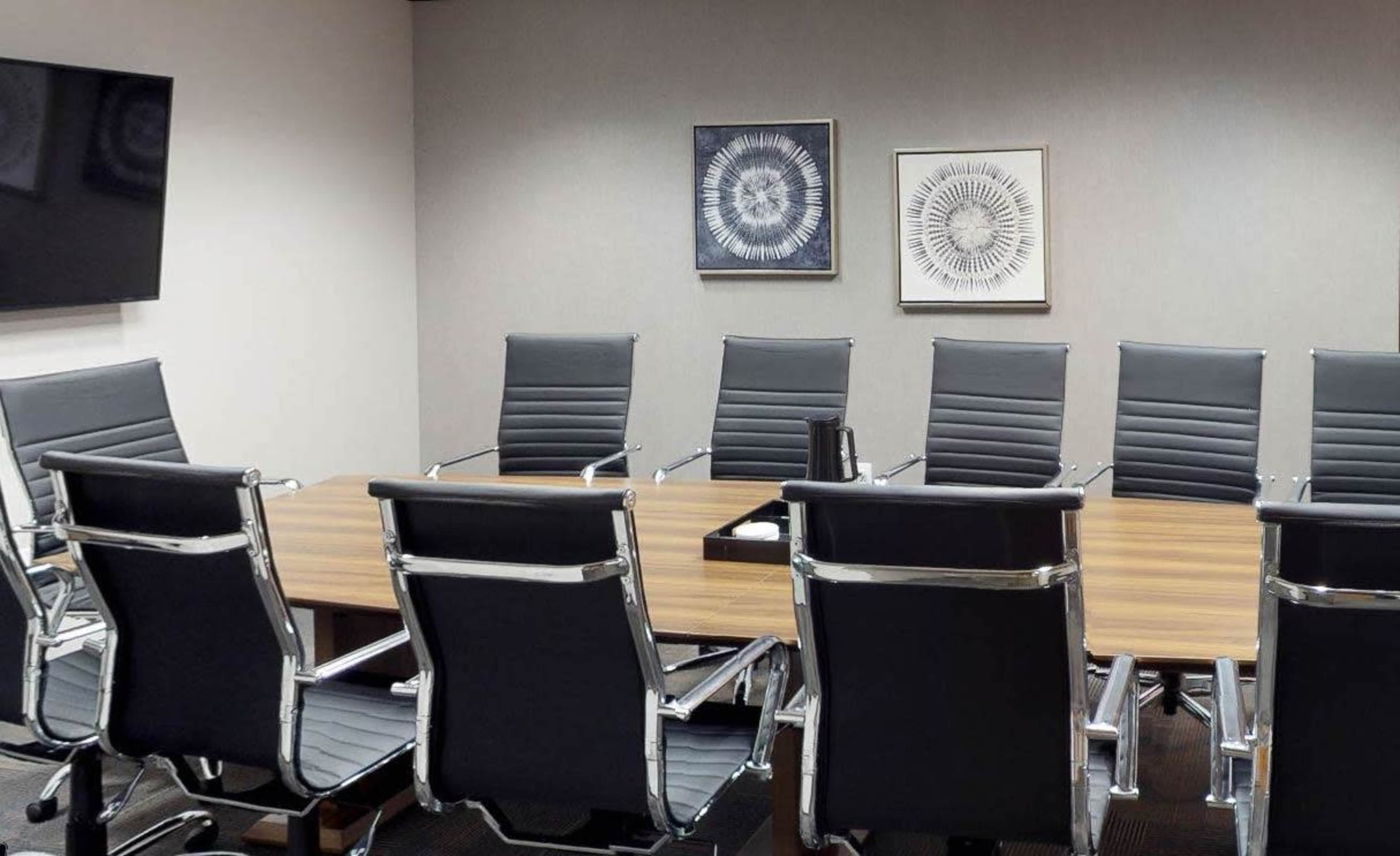 A conference room features a wooden table surrounded by eight black leather office chairs, with two framed artworks on the wall and a television mounted in the corner.
