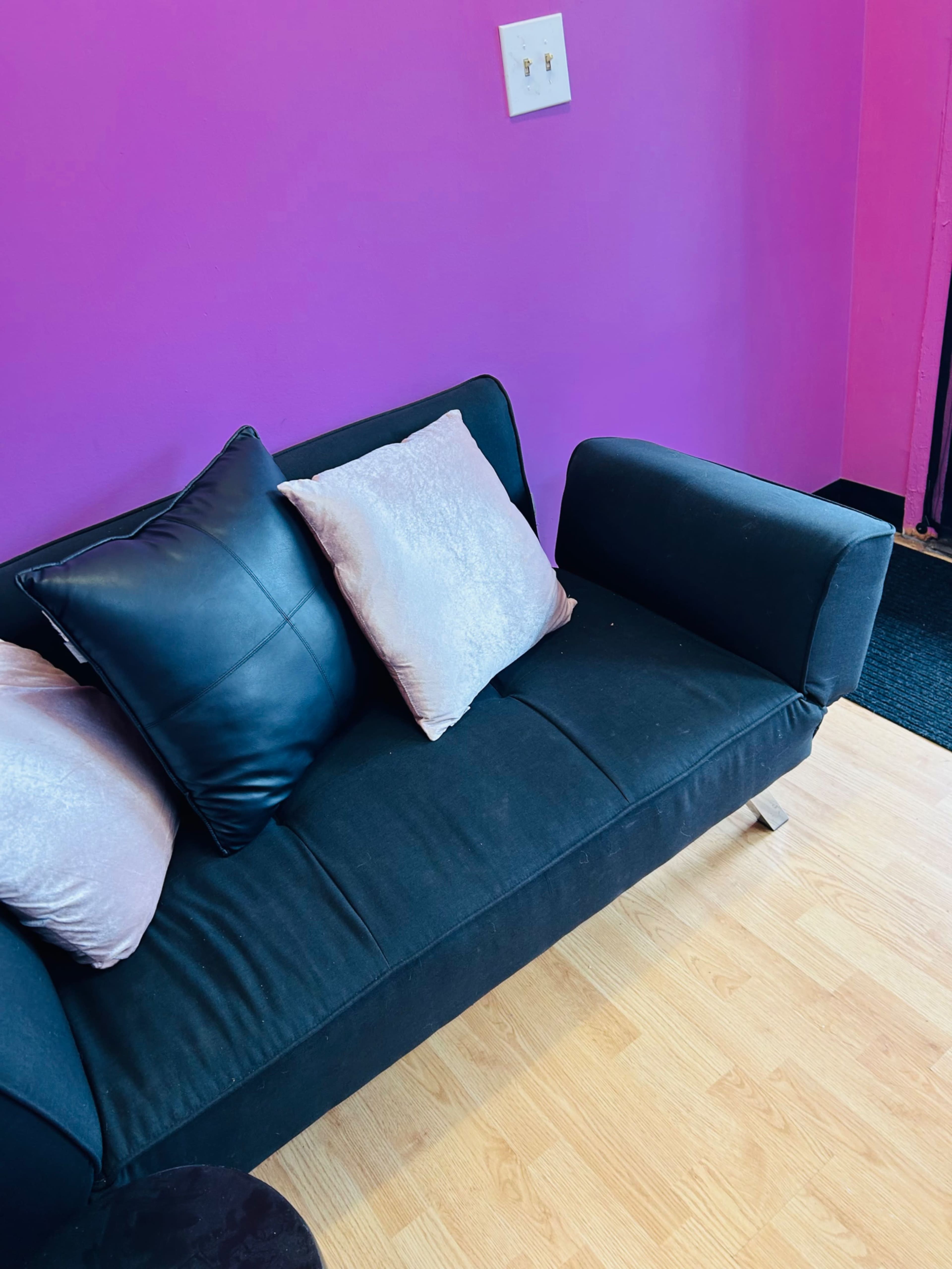 A black couch with pink pillows is positioned against a bright pink wall in a small room.