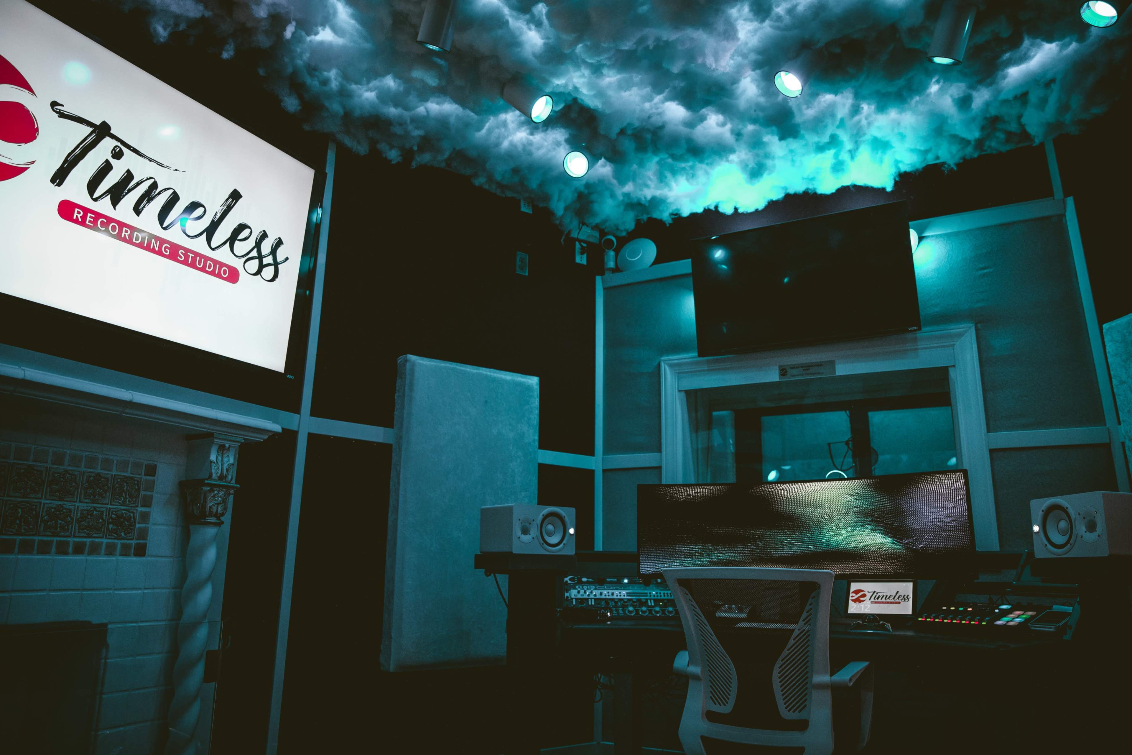 A recording studio equipped with a large mixing console, speakers, and a cloud-themed ceiling, featuring a prominent sign that says "Timeless Recording Studio."