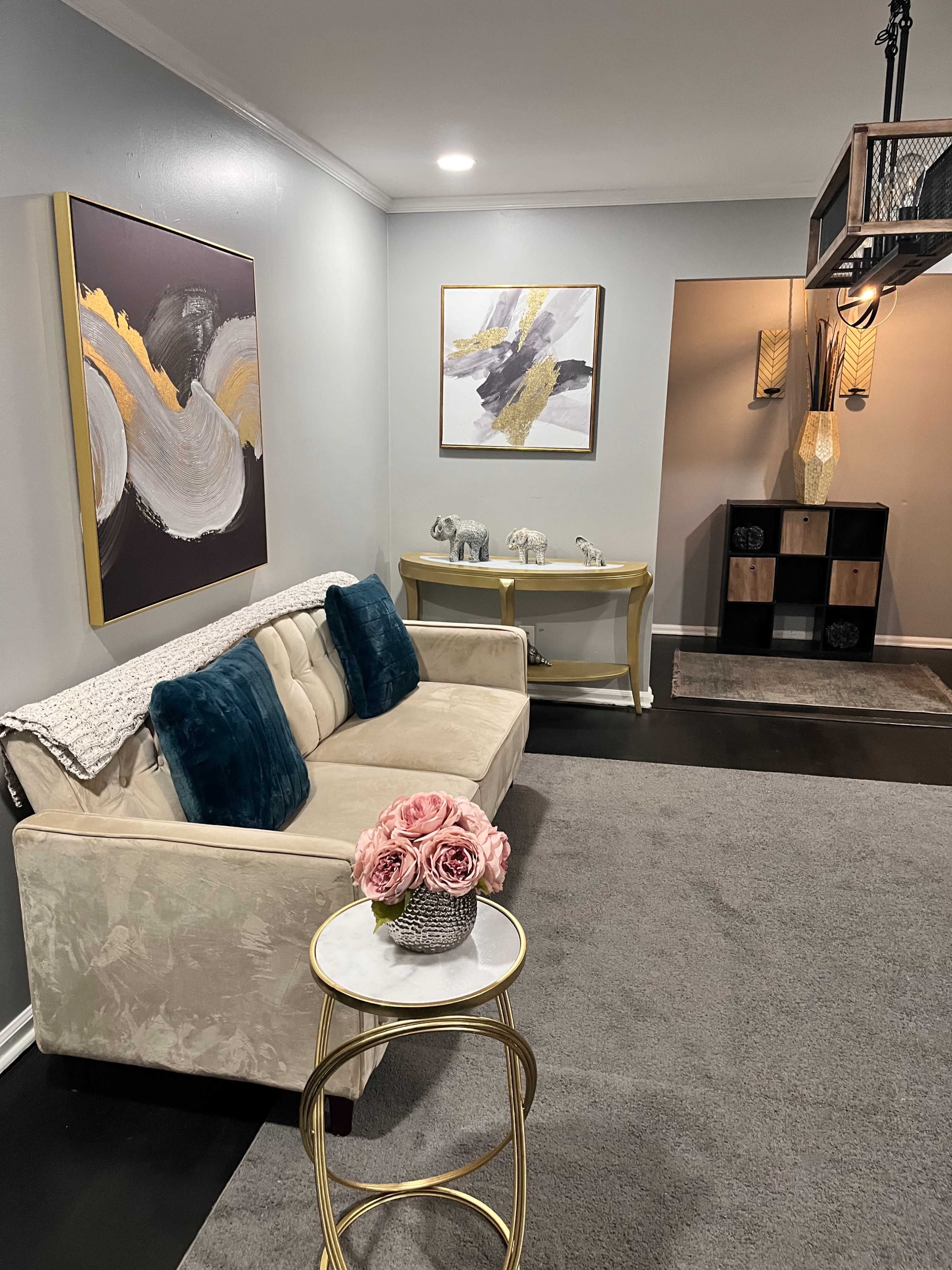 The image shows a cozy living room featuring a cream-colored sofa with teal cushions, a small round table with a floral arrangement, and two abstract paintings on the walls.