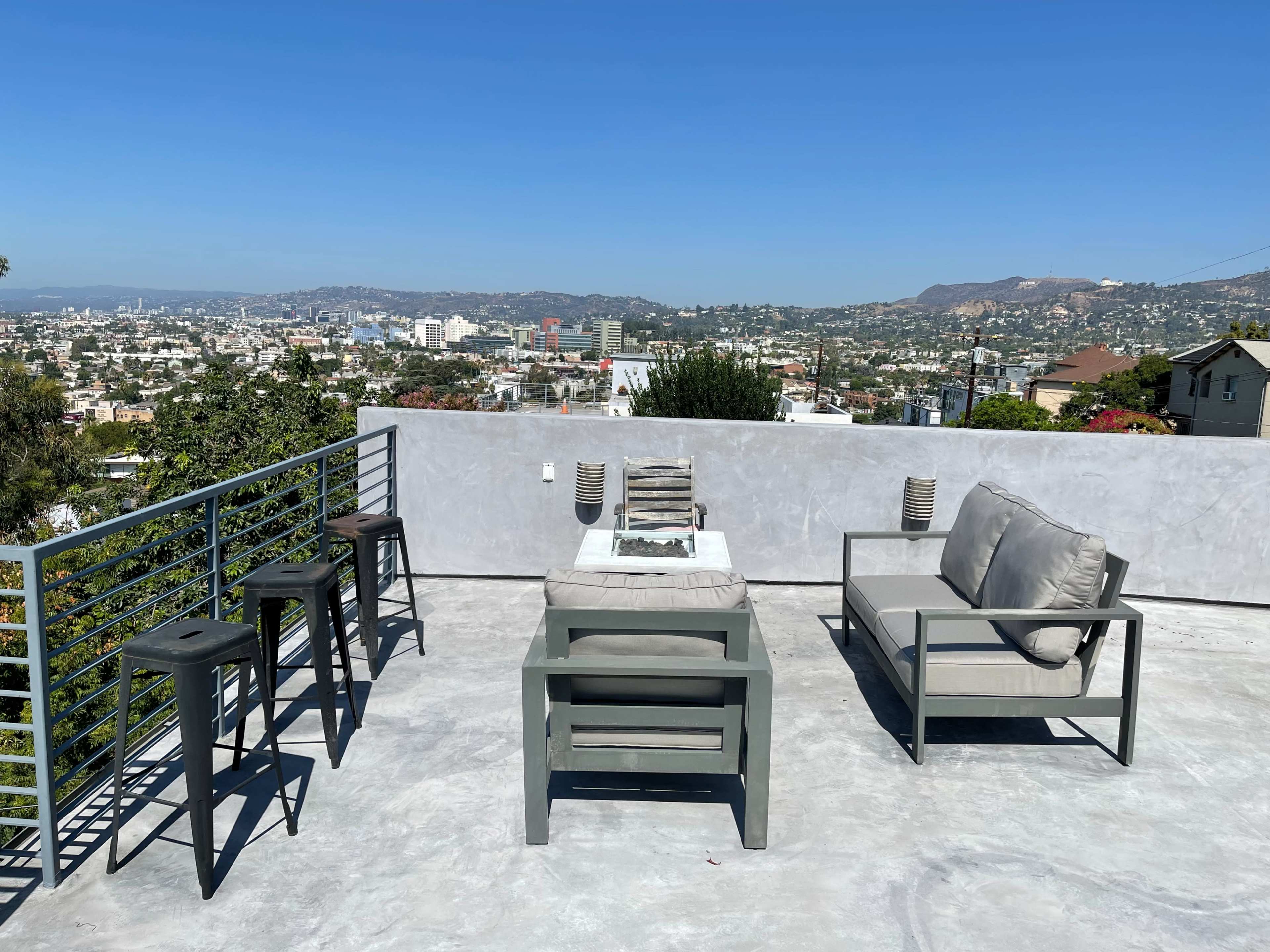 A rooftop terrace features a table with a fire pit surrounded by seating and bar stools, overlooking a cityscape.