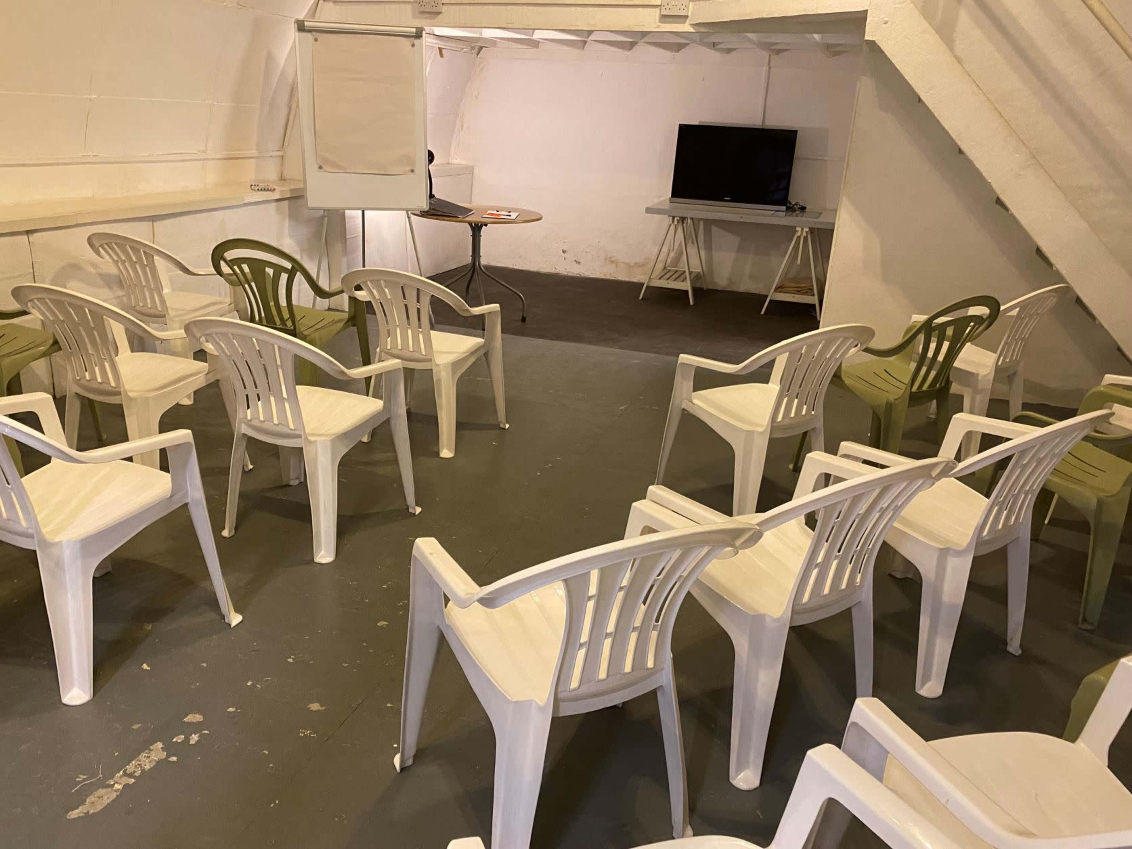 A small basement room is set up for a meeting, featuring several plastic chairs arranged in rows, a whiteboard, and a television.
