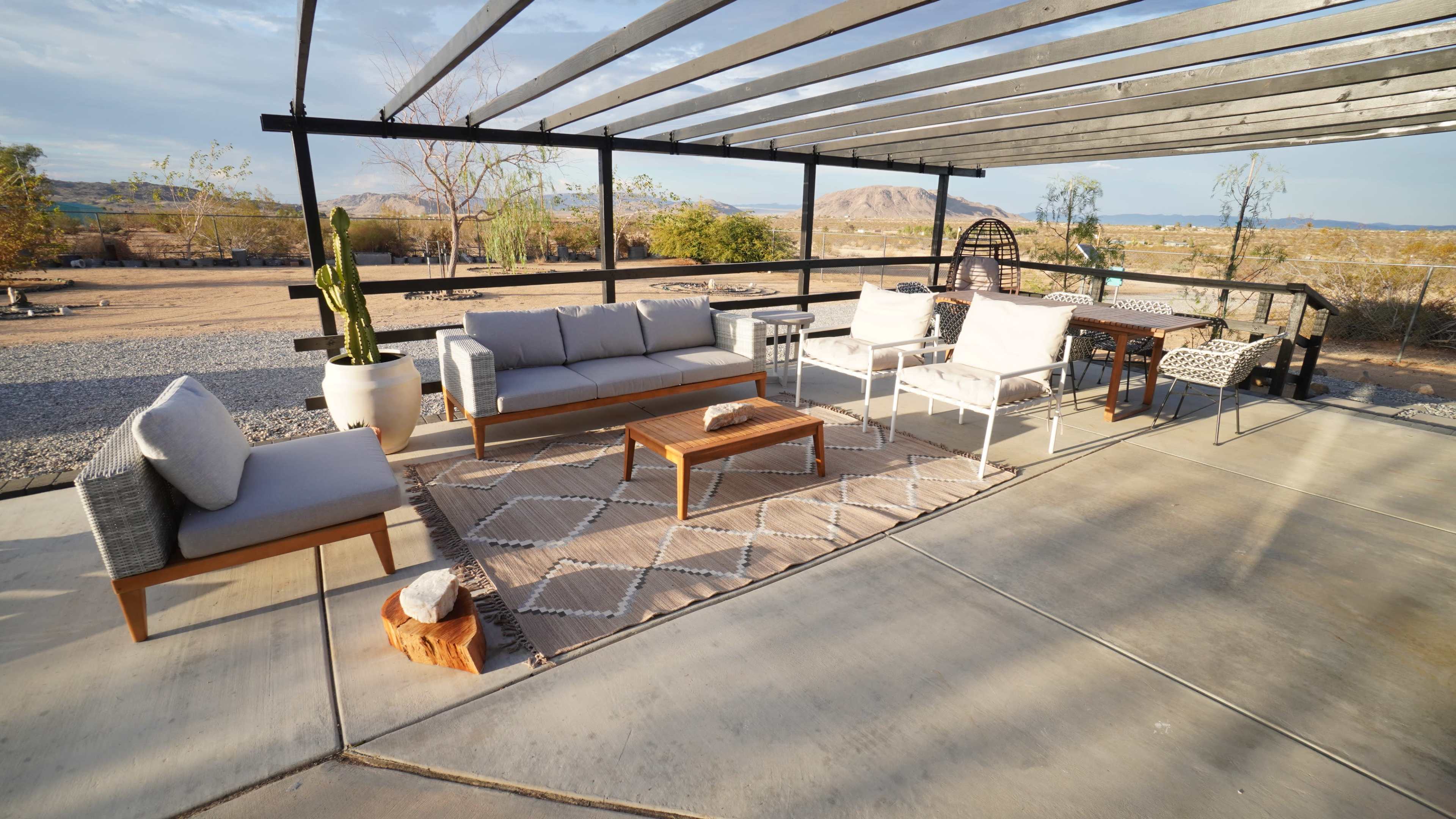 The image shows a covered outdoor seating area with modern furniture, including a sofa, chairs, and a wooden coffee table on a concrete surface, set against a desert landscape.