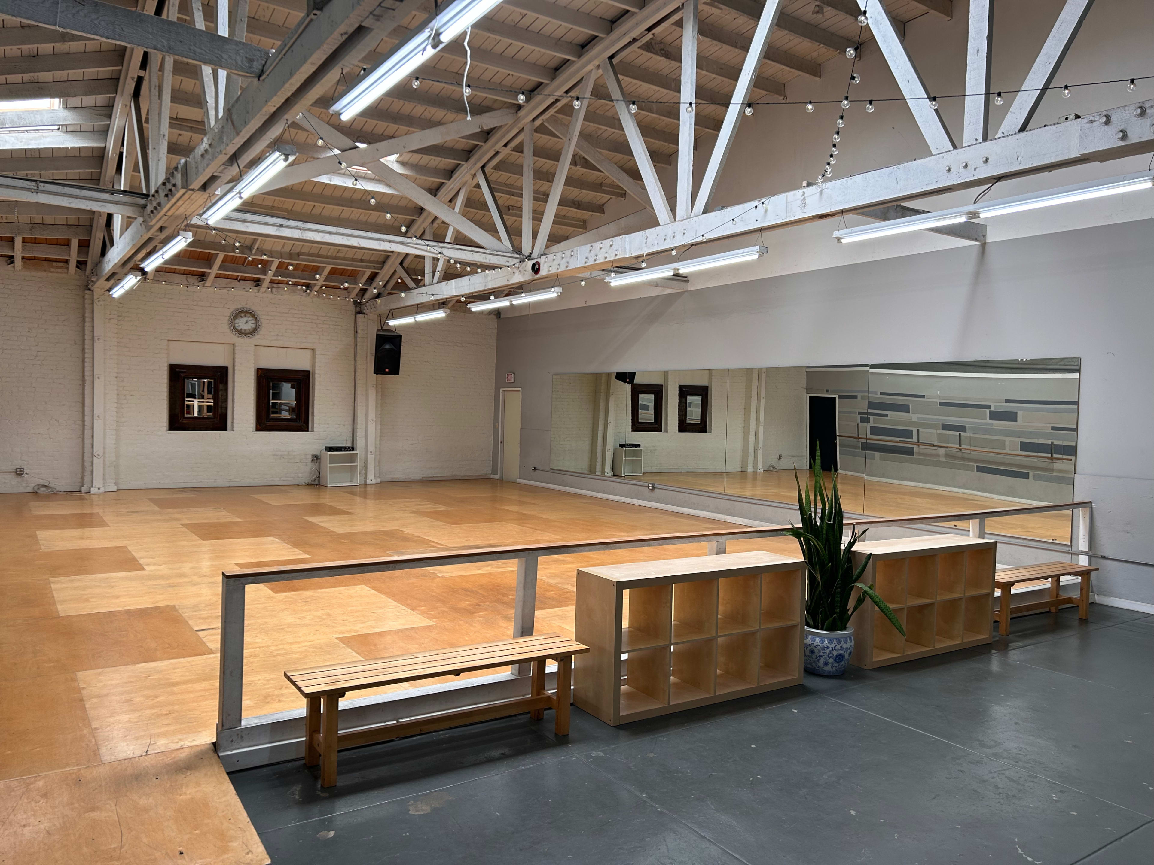 An empty dance studio with wooden flooring, mirrors on one wall, and benches along the side.
