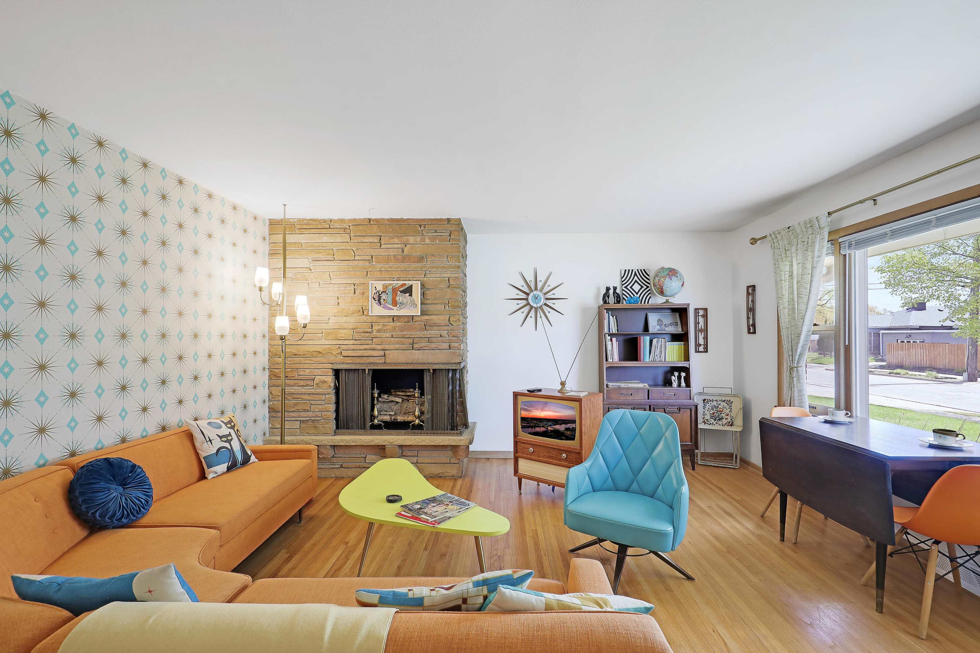 The image shows a mid-century modern living room featuring an orange sofa, a blue armchair, a stone fireplace, and stylish decorative items against a patterned wallpaper.