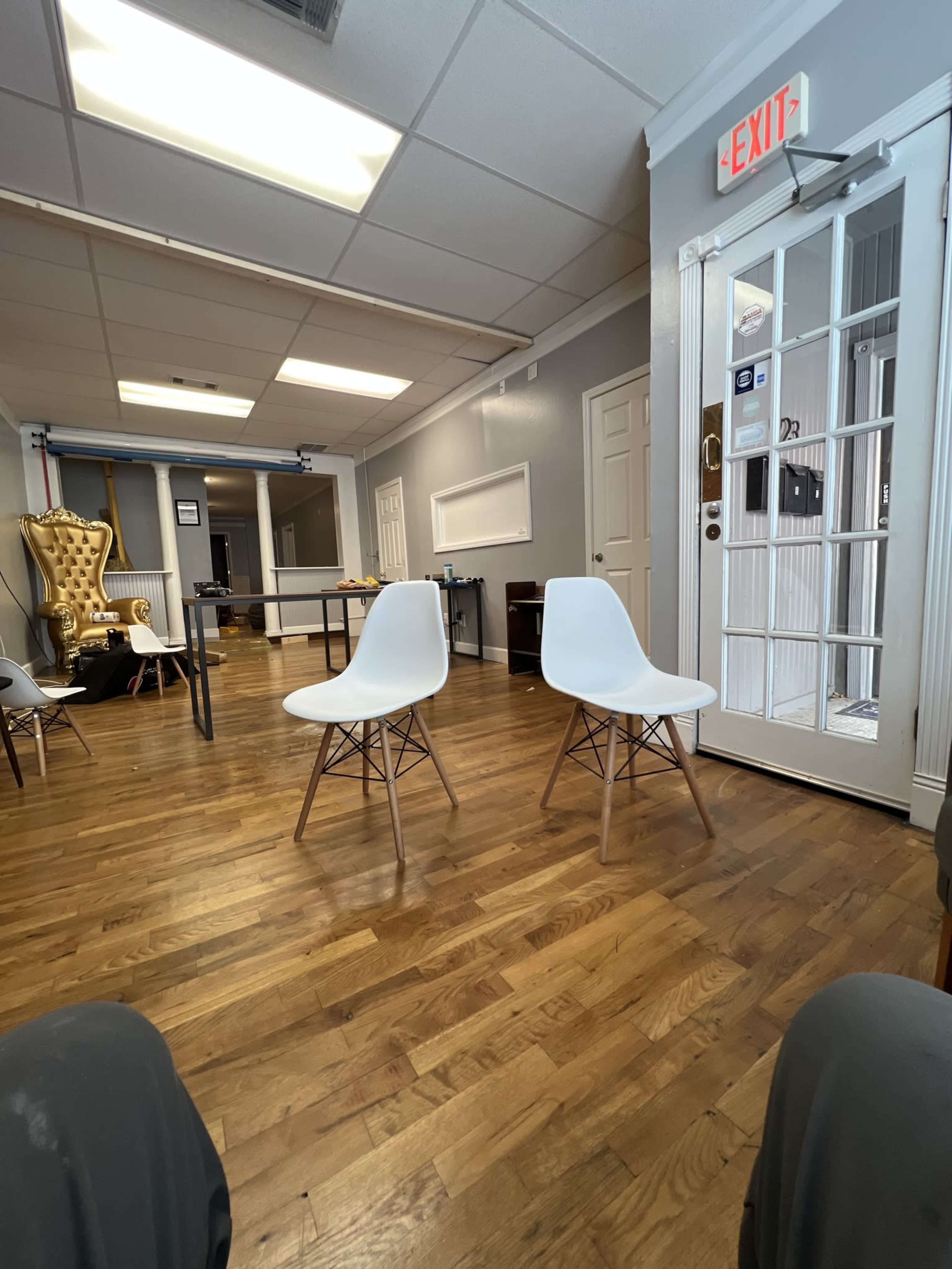 The image shows a spacious, empty room with two white chairs, a table in the center, and a door leading outside.