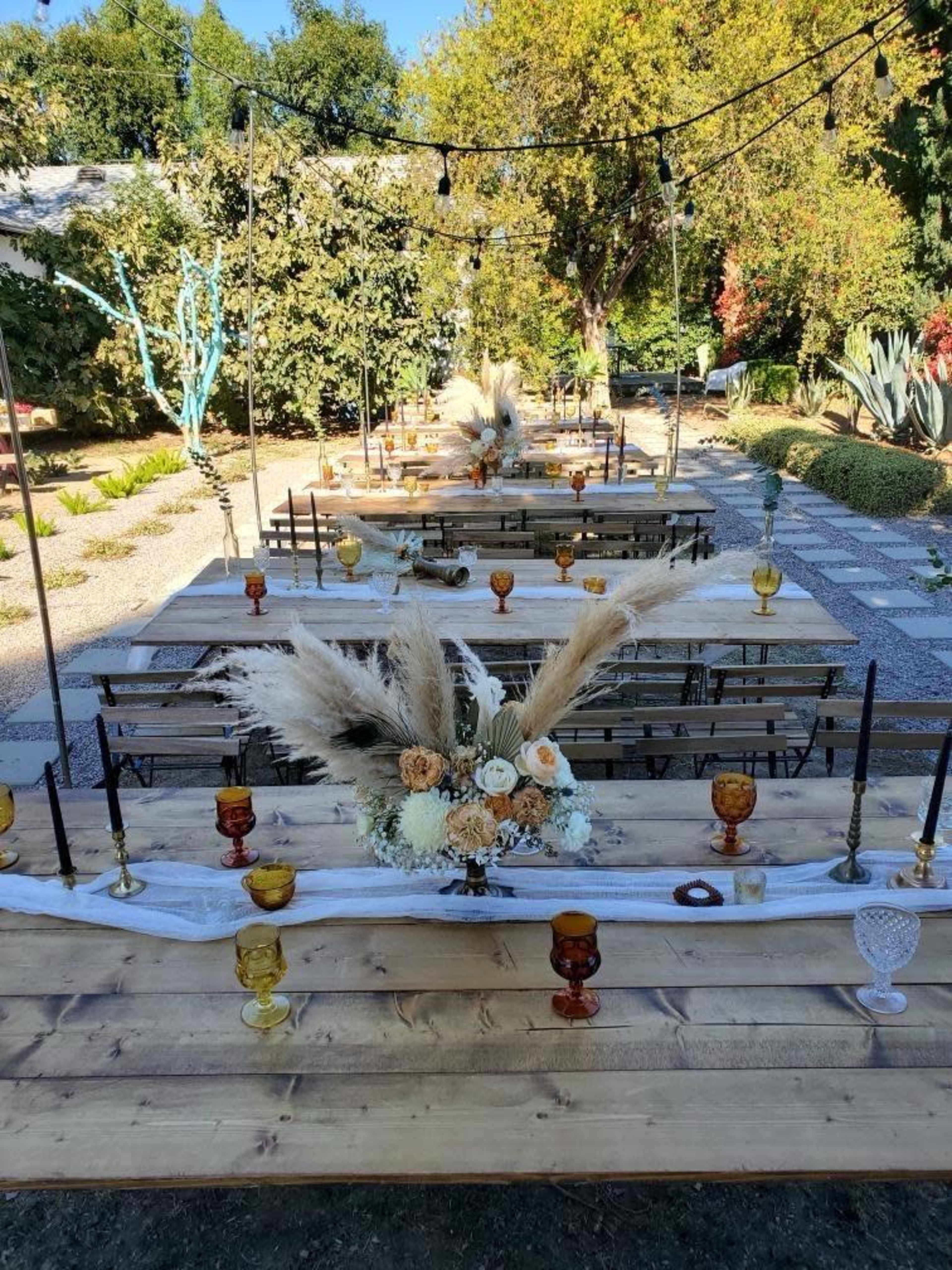 A beautifully arranged outdoor dining setup with long wooden tables, adorned with floral centerpieces and decorative candles, in a garden setting.