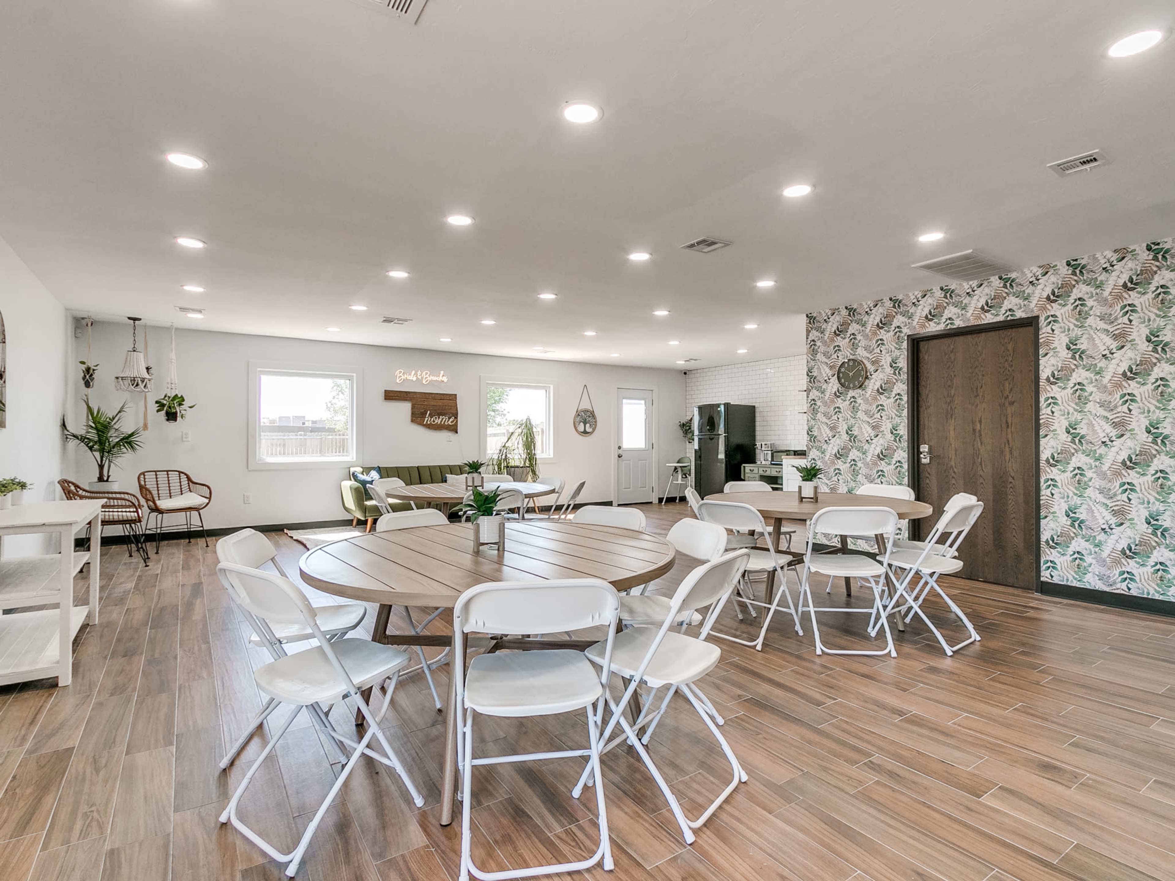 A spacious interior featuring multiple round tables with white chairs, a green sofa, and a modern kitchen area against a patterned wall.