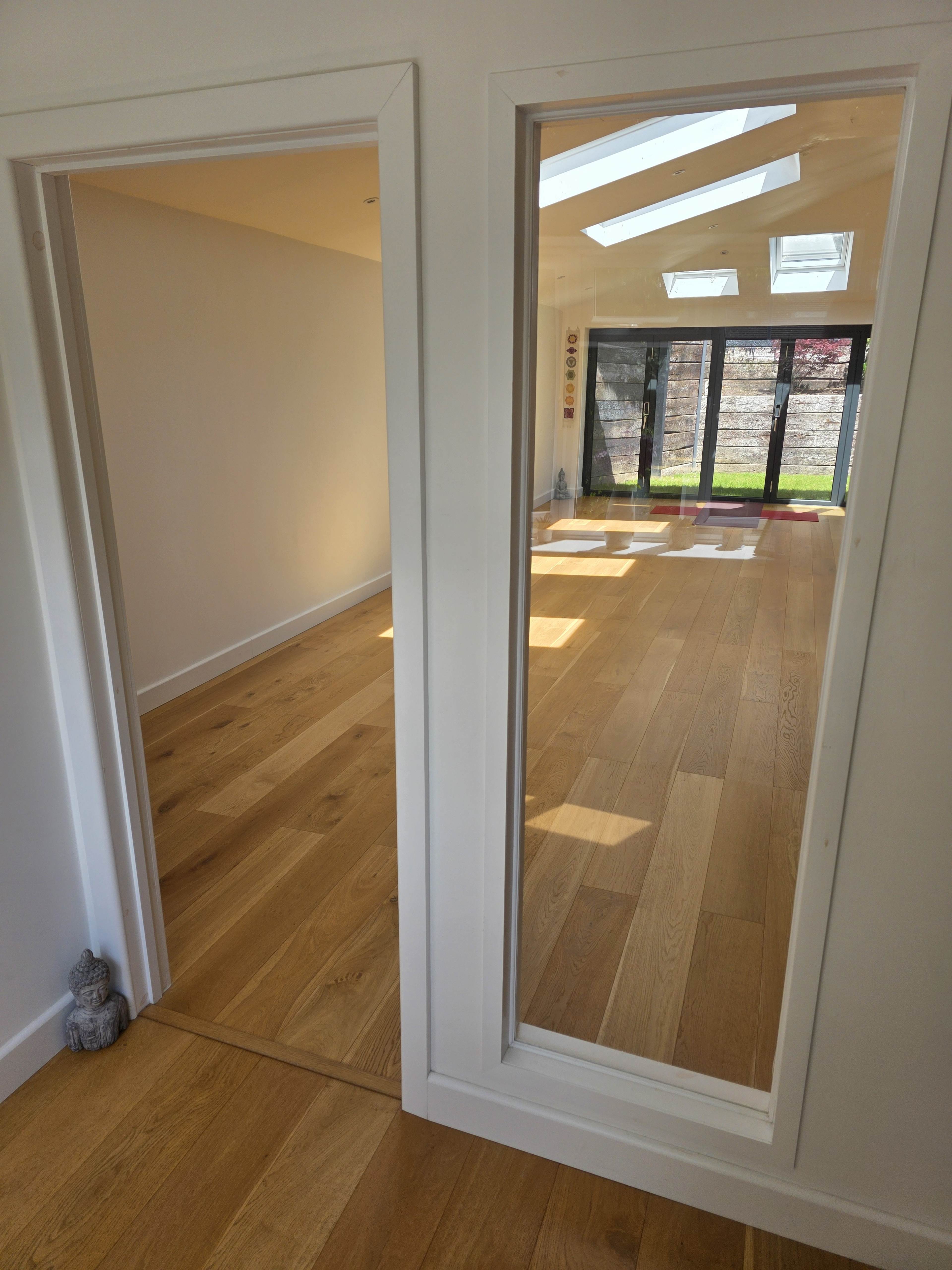 The image shows a hallway with a mirror reflecting a room that has large windows and wooden flooring.