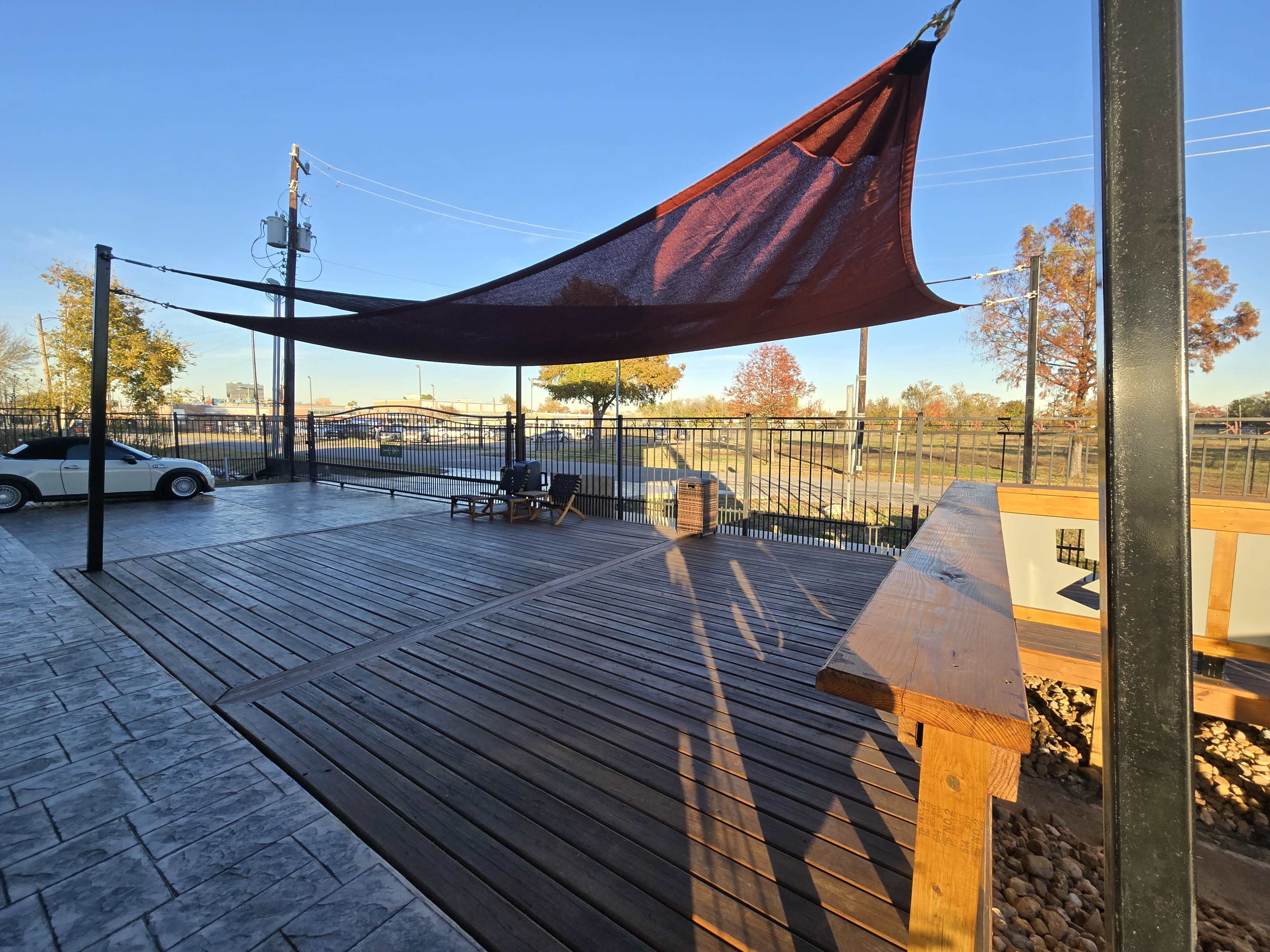 A shaded wooden deck with a maroon canopy overlooks a fenced area, featuring a car and a grassy field in the background.