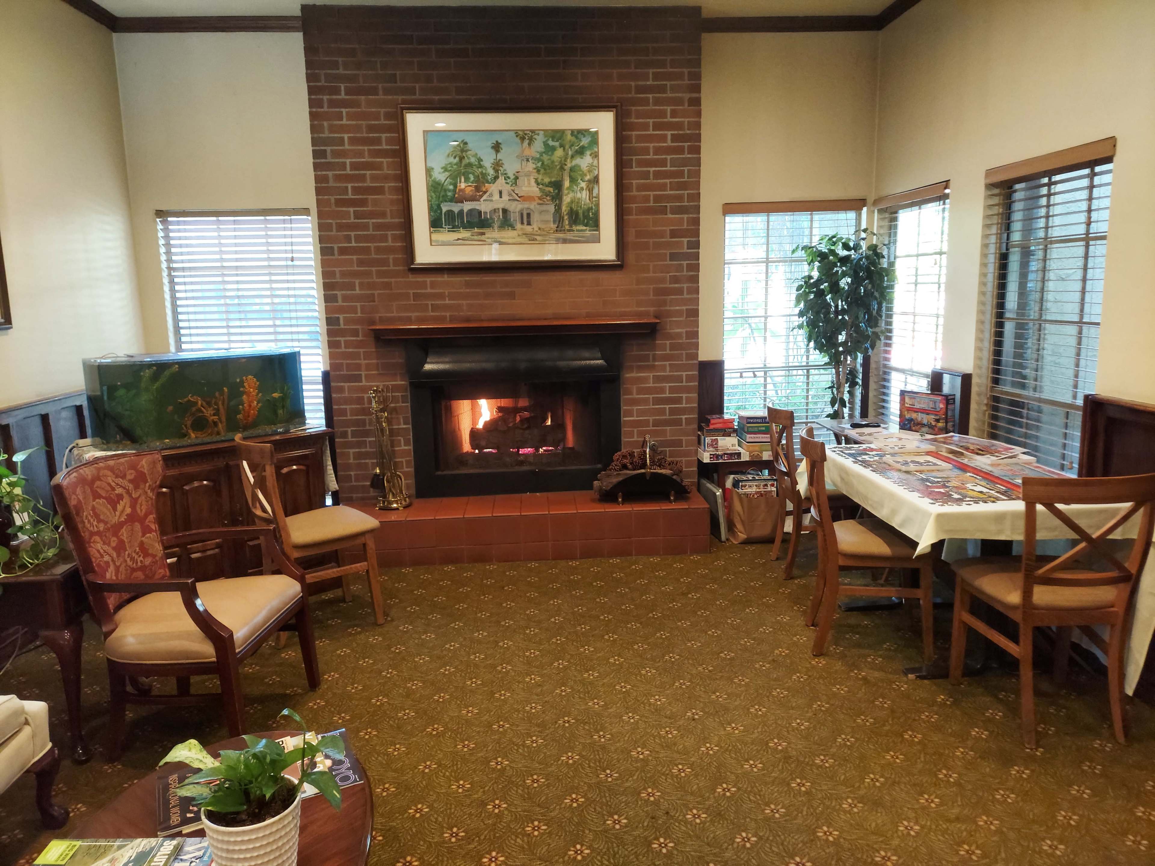 The image shows a cozy living room with a fireplace, a fish tank, seating arrangements, and a table surrounded by chairs.