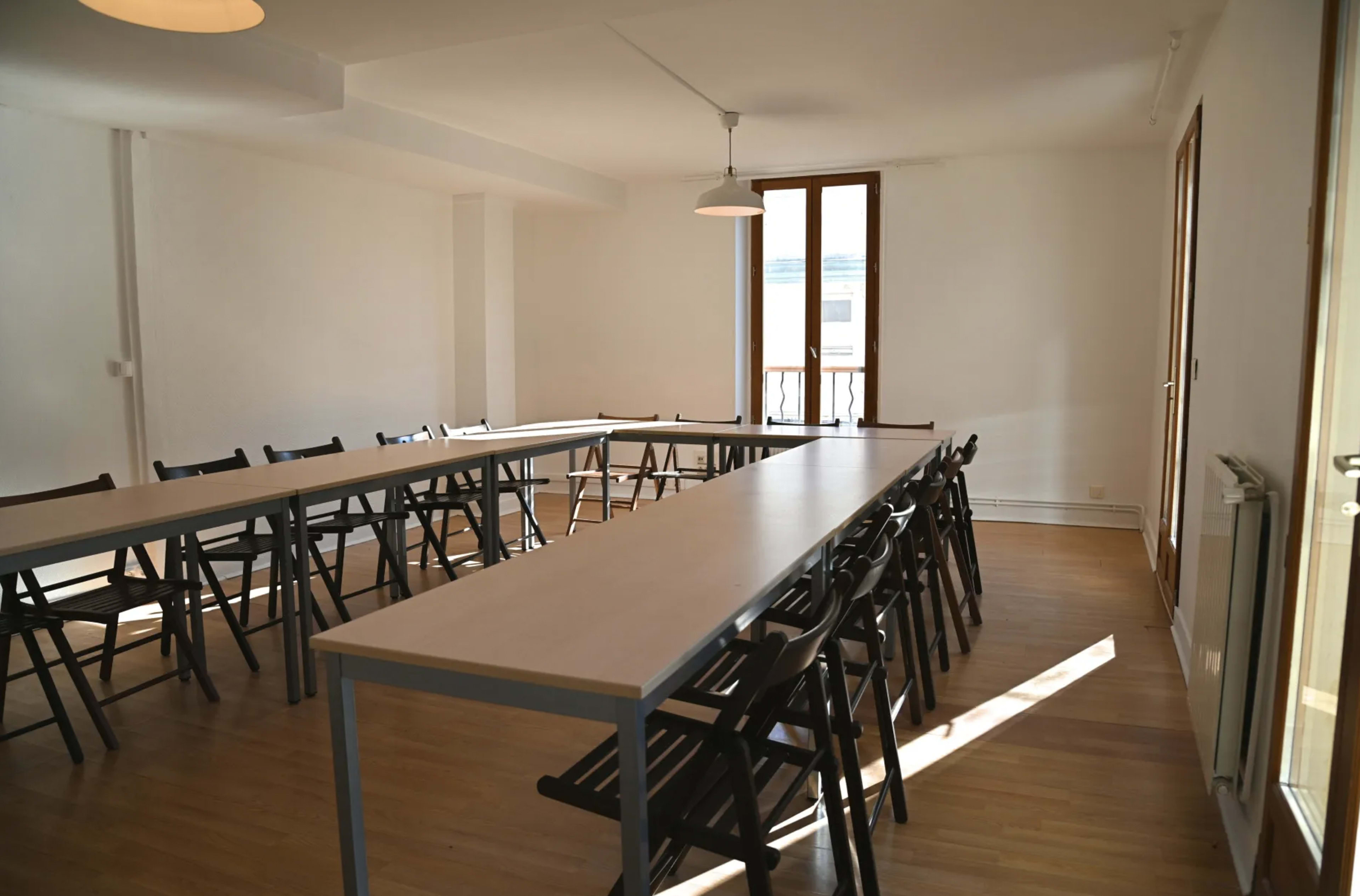 A spacious meeting room features several long tables arranged in a U-shape with black chairs around them and large windows allowing natural light.