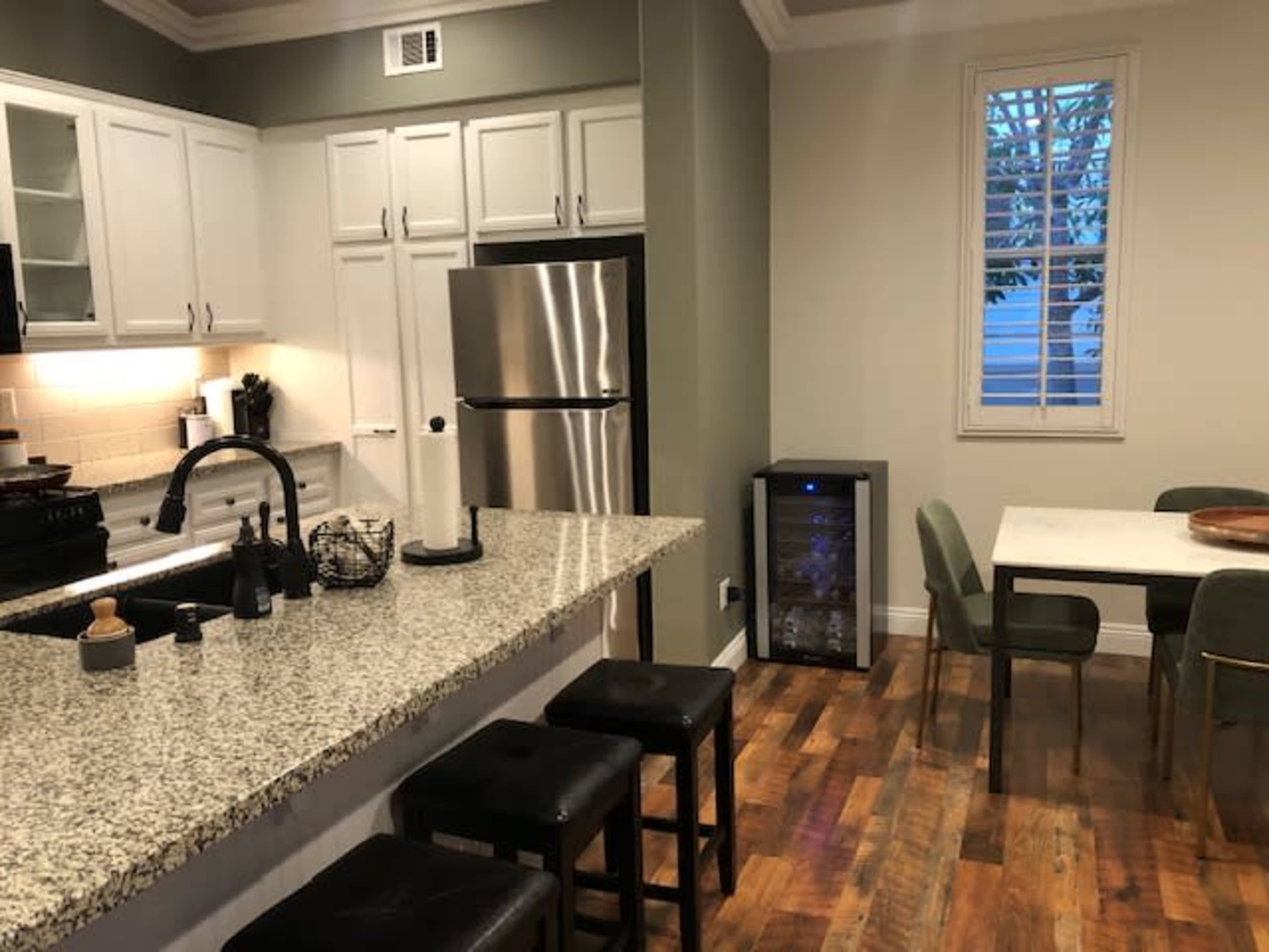 The image shows a modern kitchen with white cabinetry, stainless steel appliances, a granite countertop, and a dining table with green chairs.