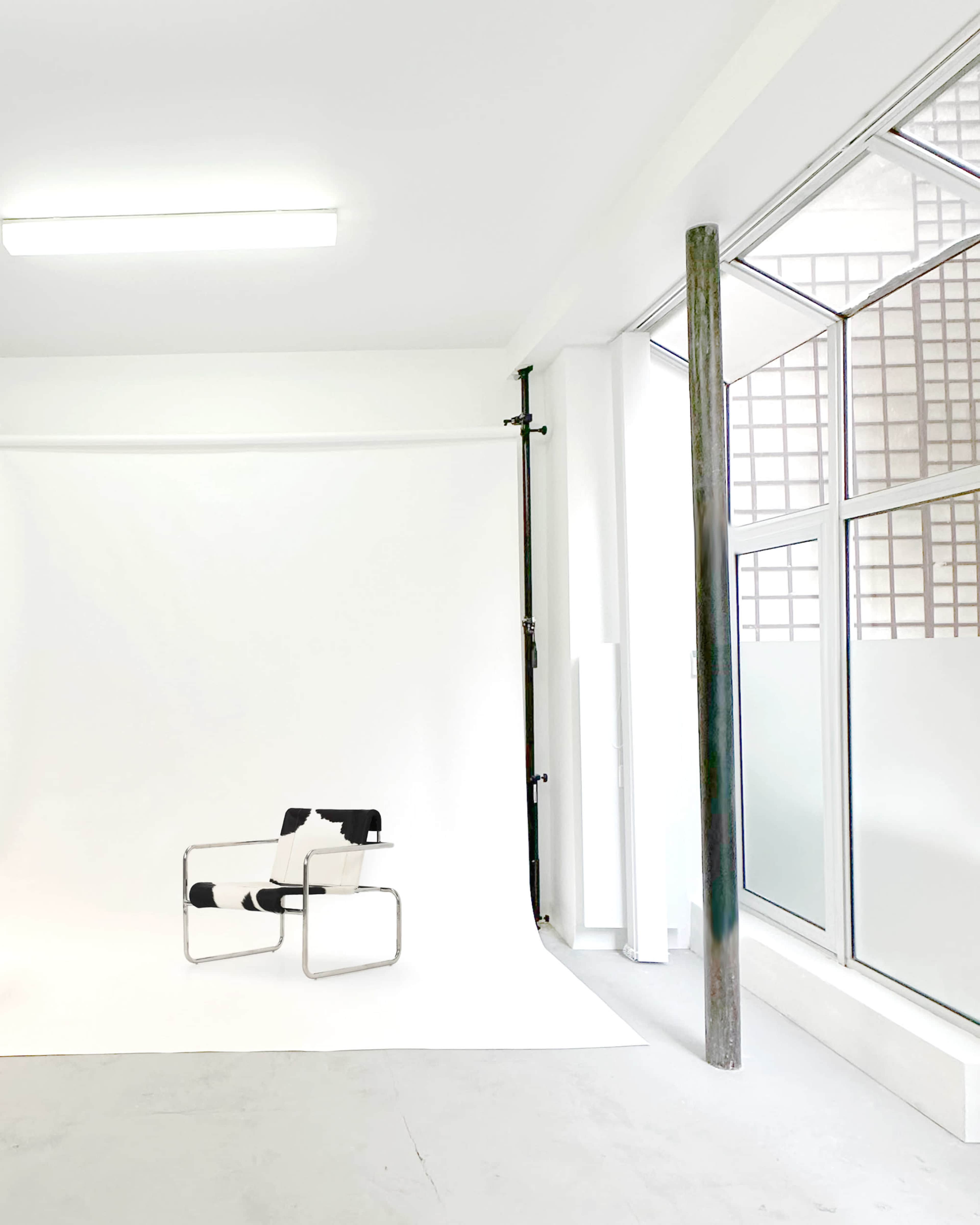 The image shows a minimalist room with a black and white patterned chair on a white backdrop and large windows allowing natural light to enter.
