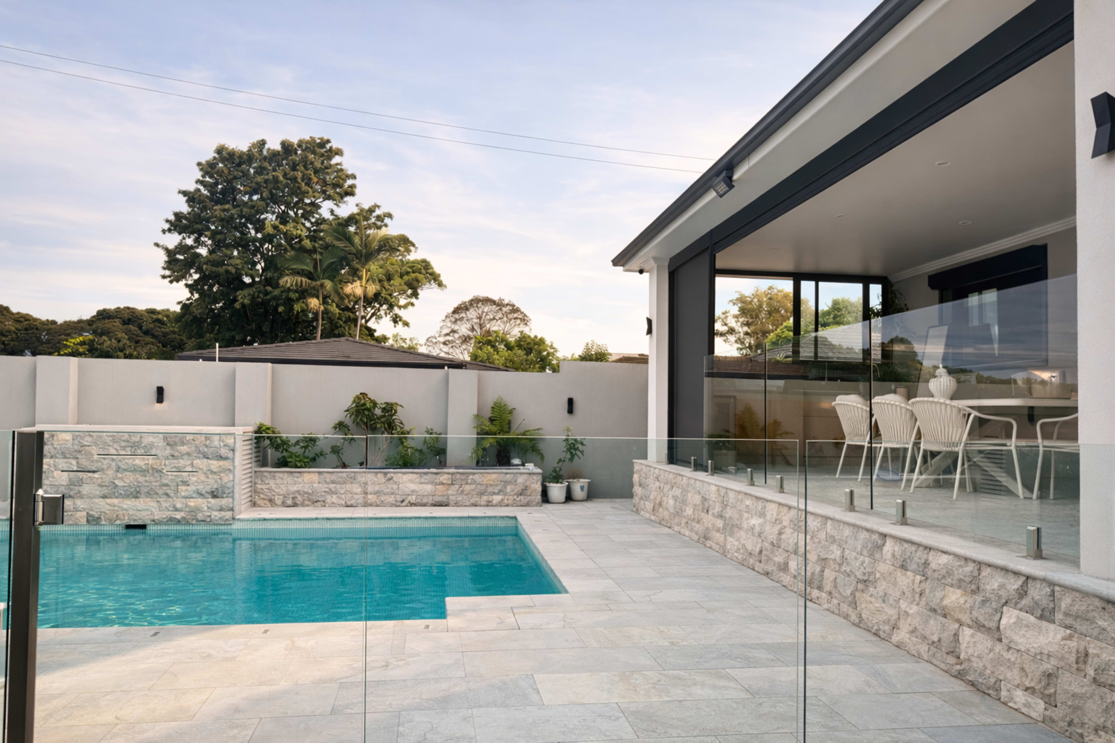 A modern outdoor space features a swimming pool, stone patio, and a covered dining area with glass railings.