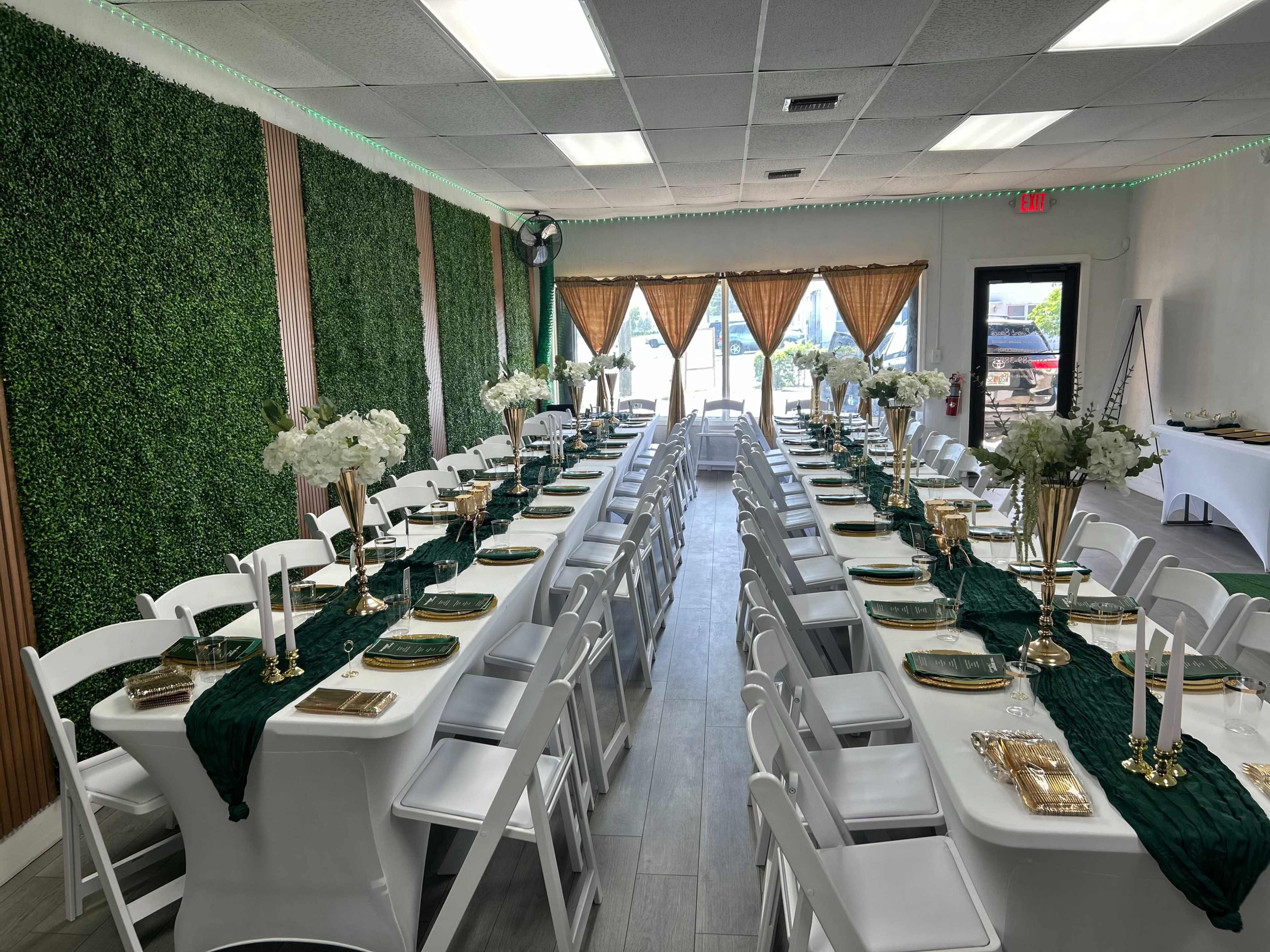 The image shows a beautifully arranged event space with long dining tables set with green tablecloths, elegant floral centerpieces, and golden tableware, surrounded by greenery-covered walls and large windows.