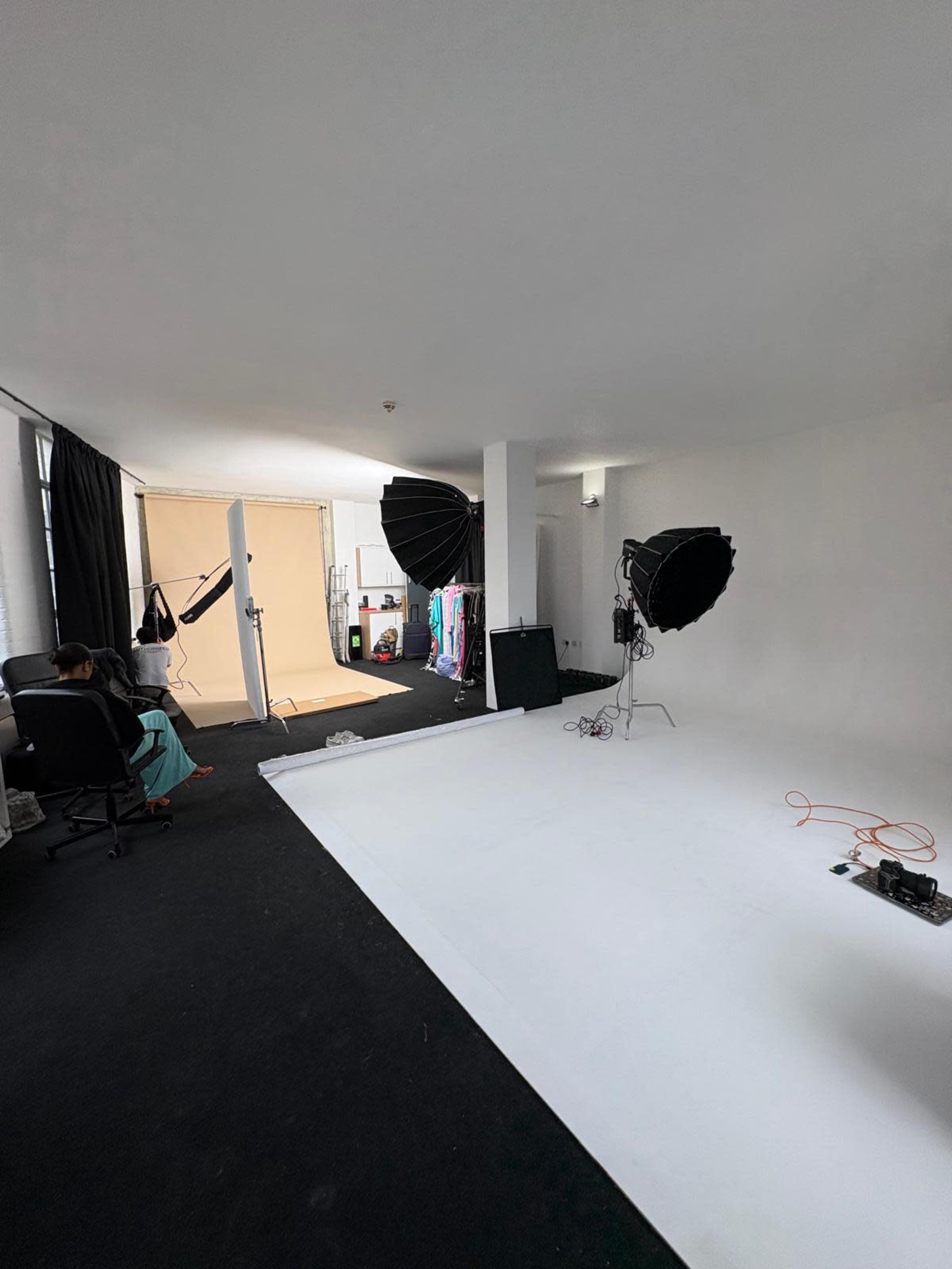 The image shows a photography studio featuring two large softbox lights, a backdrop area with a light-colored floor, and various props and clothing in the background.