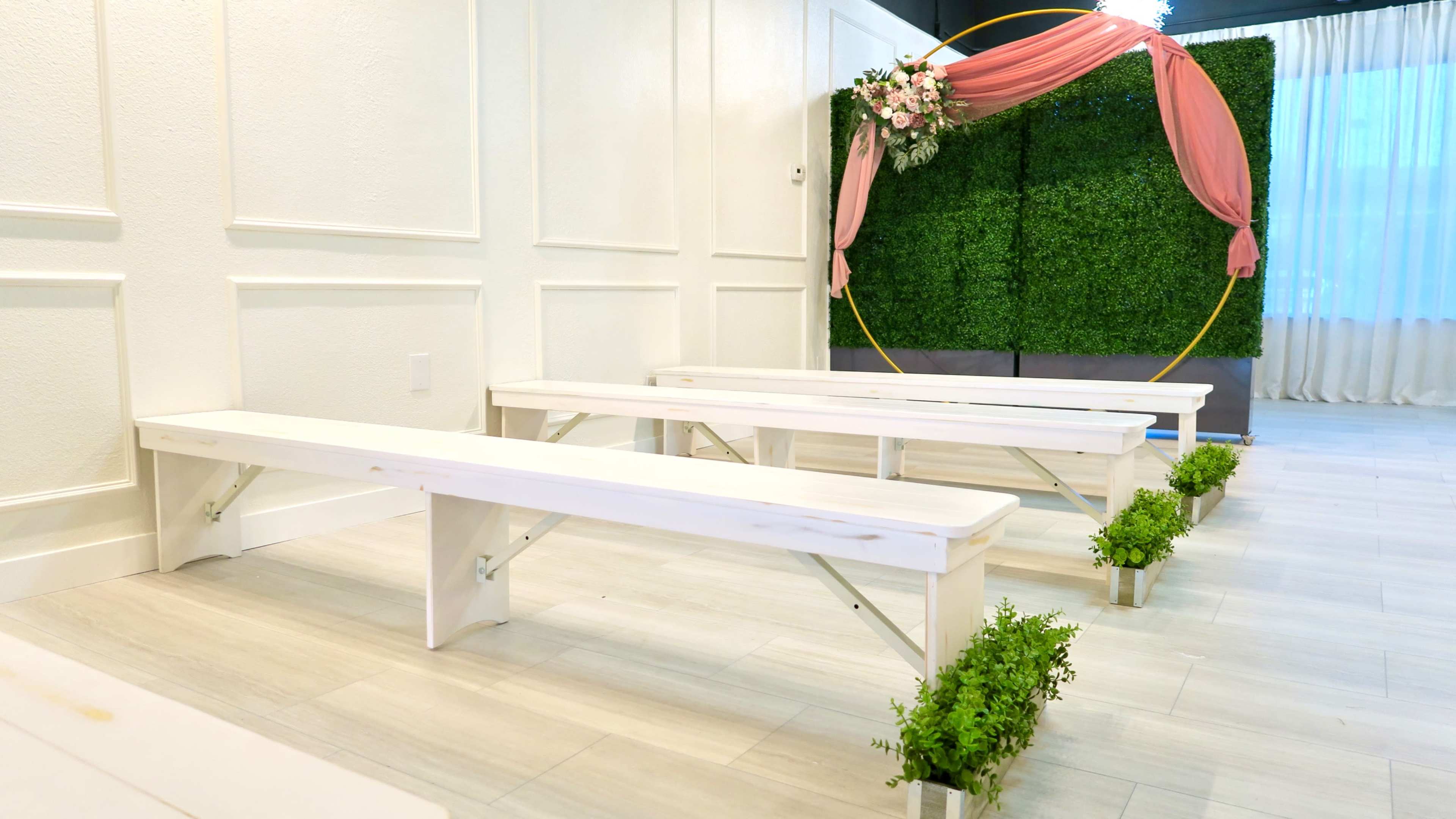 The image shows a simple event setup with white benches arranged on a light-colored floor facing a decorative arch covered with greenery and a pink drape.