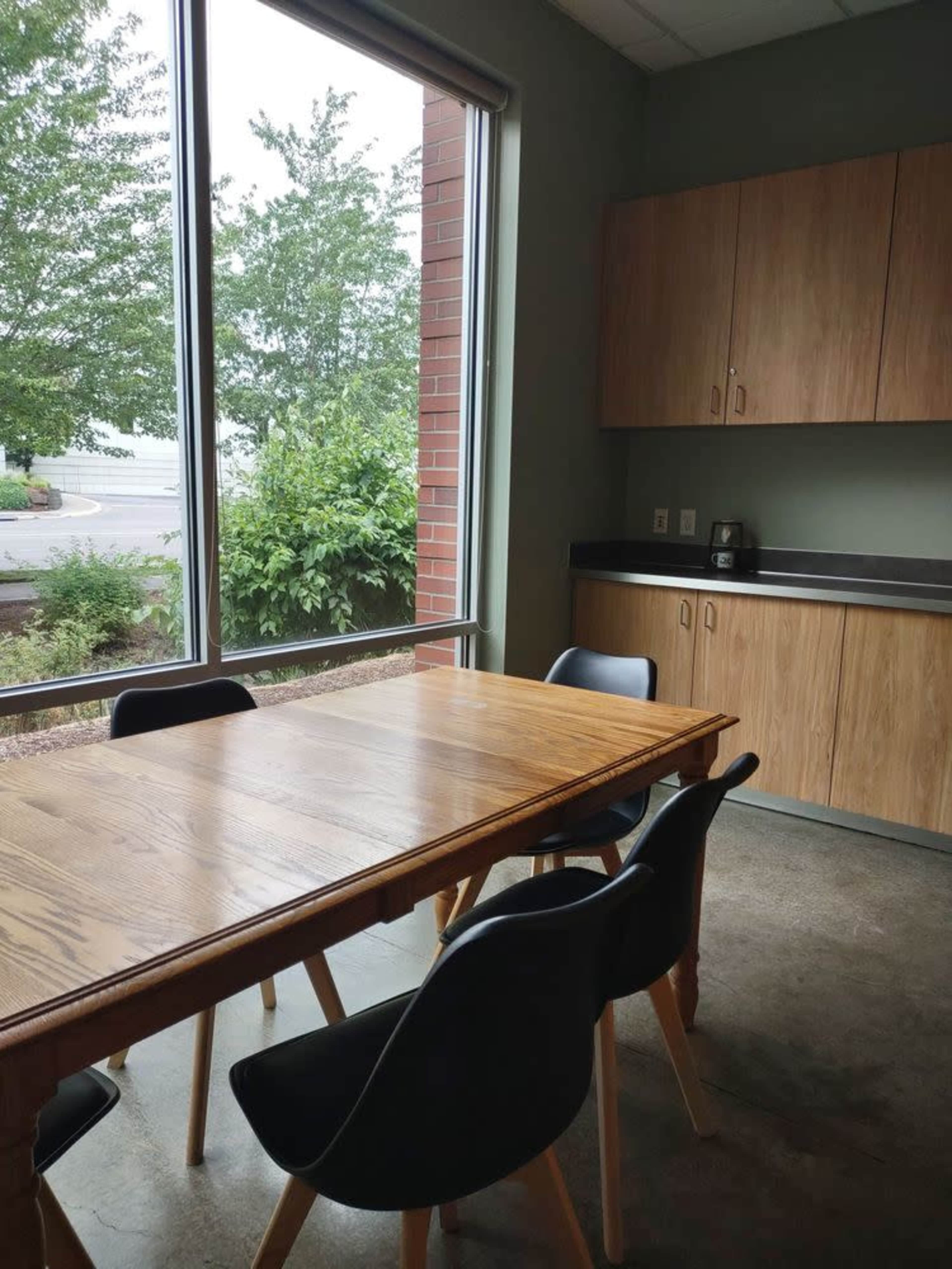 A wooden table with black chairs is positioned near a large window beside a kitchenette with upper cabinets and a coffee maker.