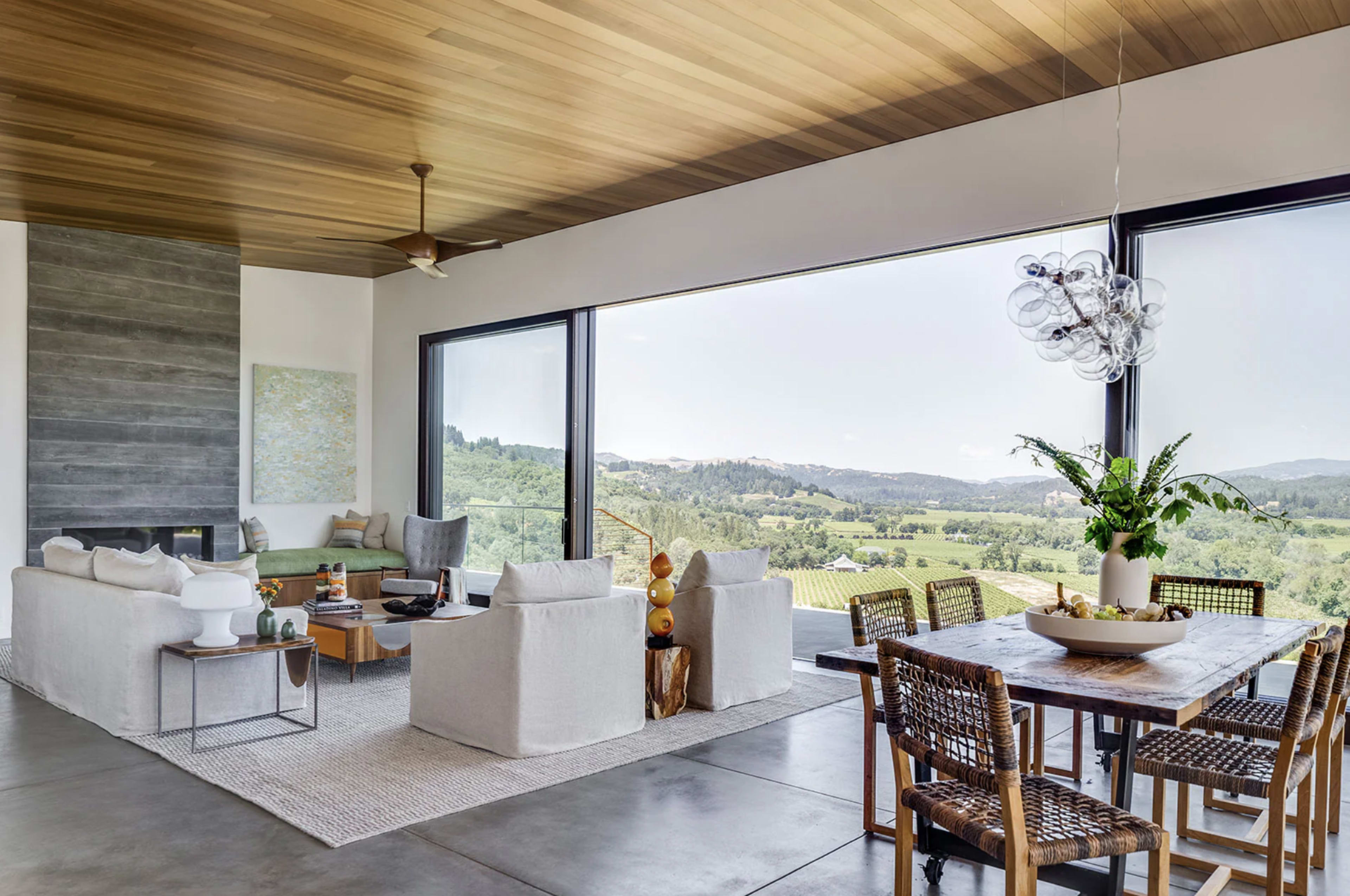 A modern living room features large windows with a scenic view, comfortable seating, and a dining area with a wooden table surrounded by woven chairs.