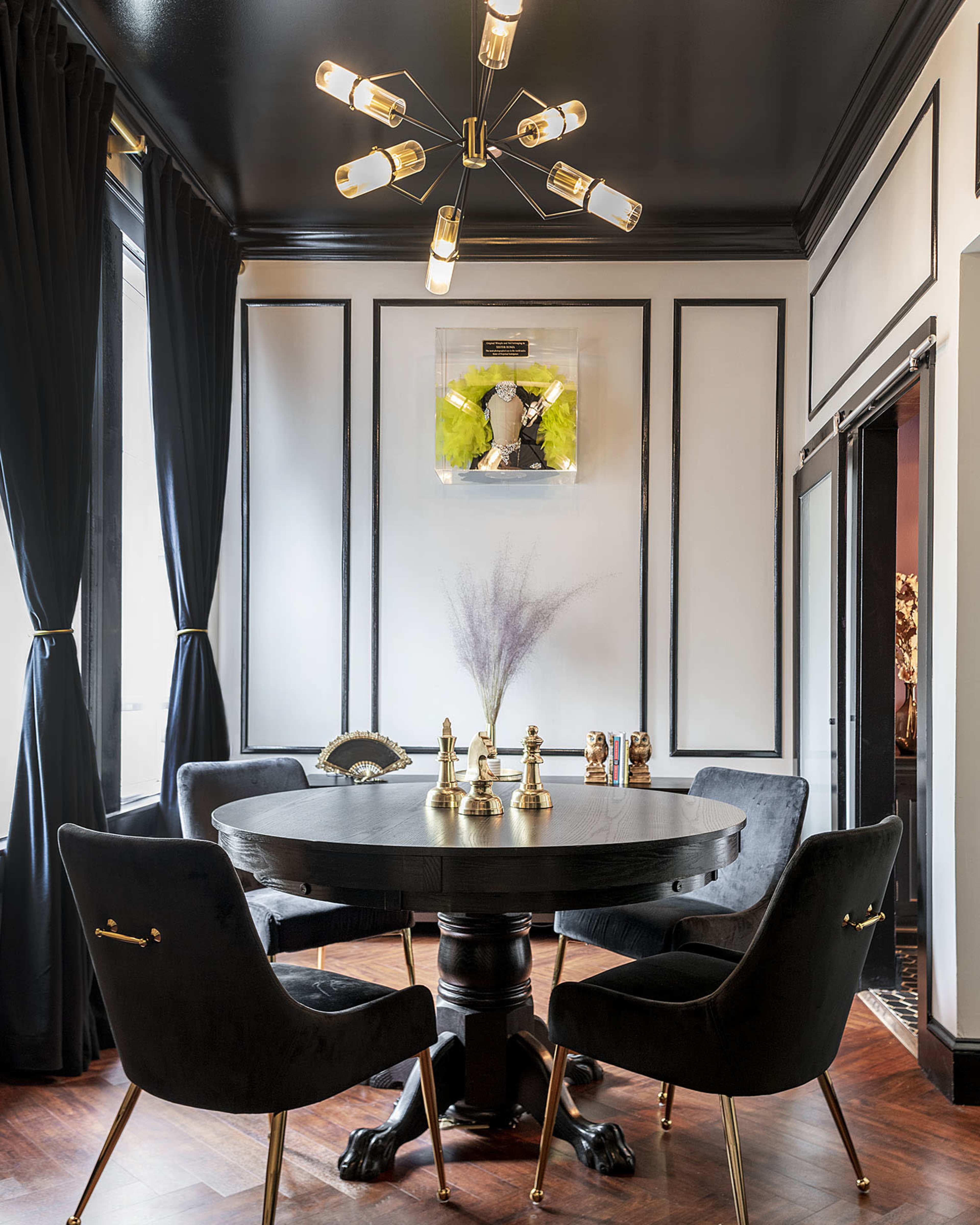 A dimly lit dining area featuring a round black table surrounded by two upholstered chairs, adorned with decorative objects on the table and framed artwork on the wall.