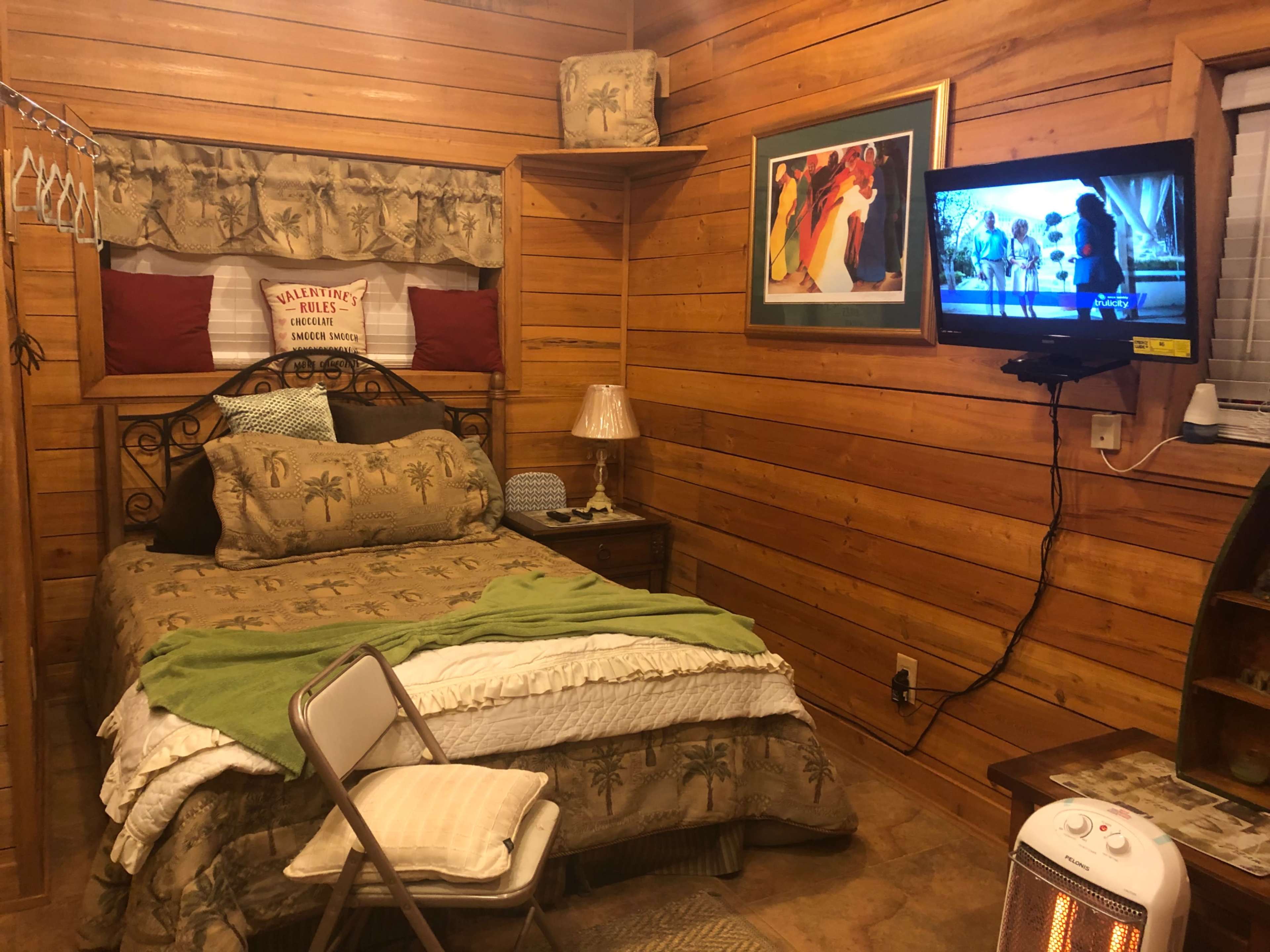 A cozy wooden bedroom featuring a bed with tropical-themed linens, a small bedside lamp, a television mounted on the wall, and a folding chair.