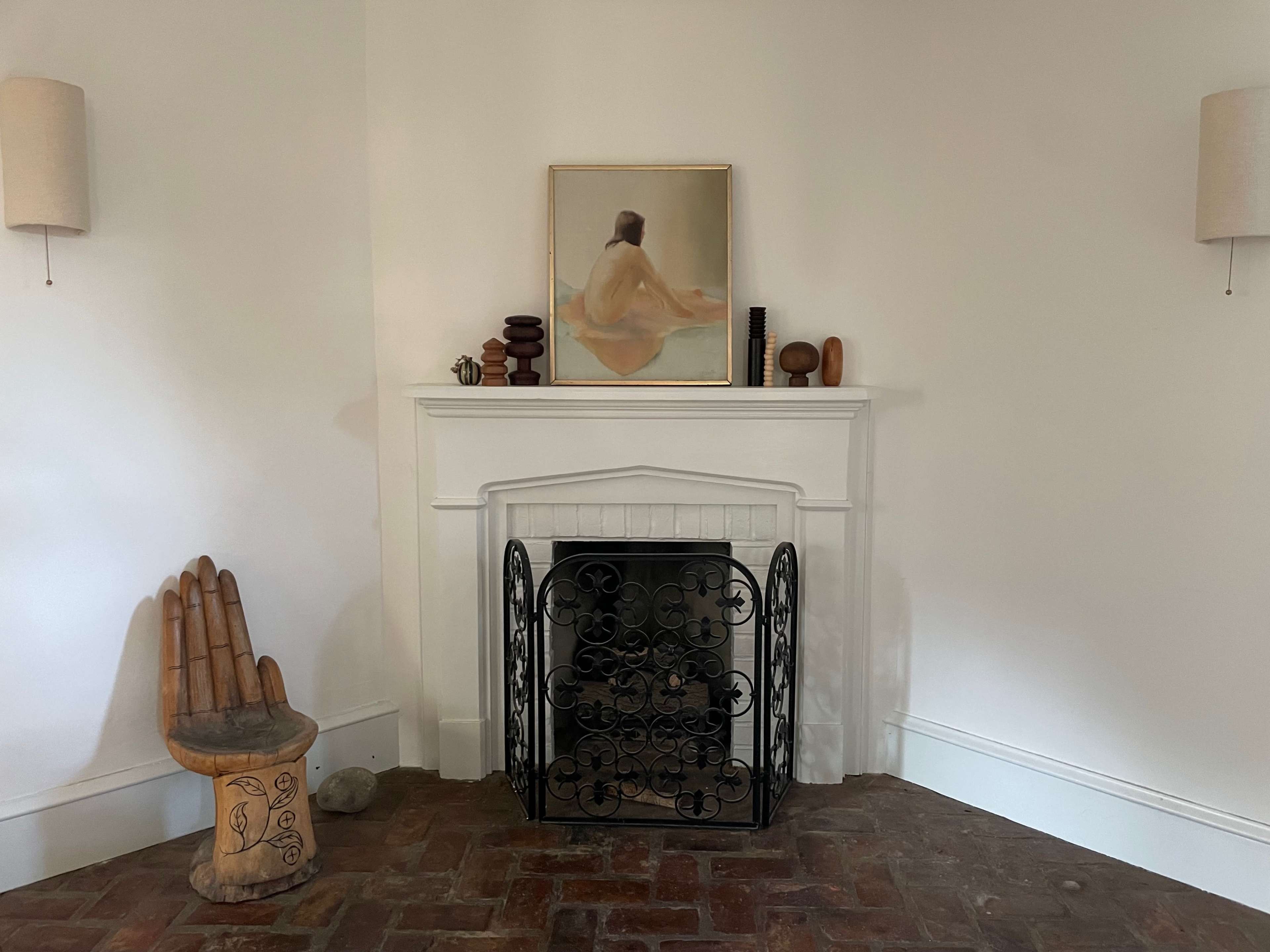 A simple room with a brick floor, featuring a fireplace with a black wrought iron screen, an artwork above it, and several decorative objects, including a carved wooden stool shaped like a hand.