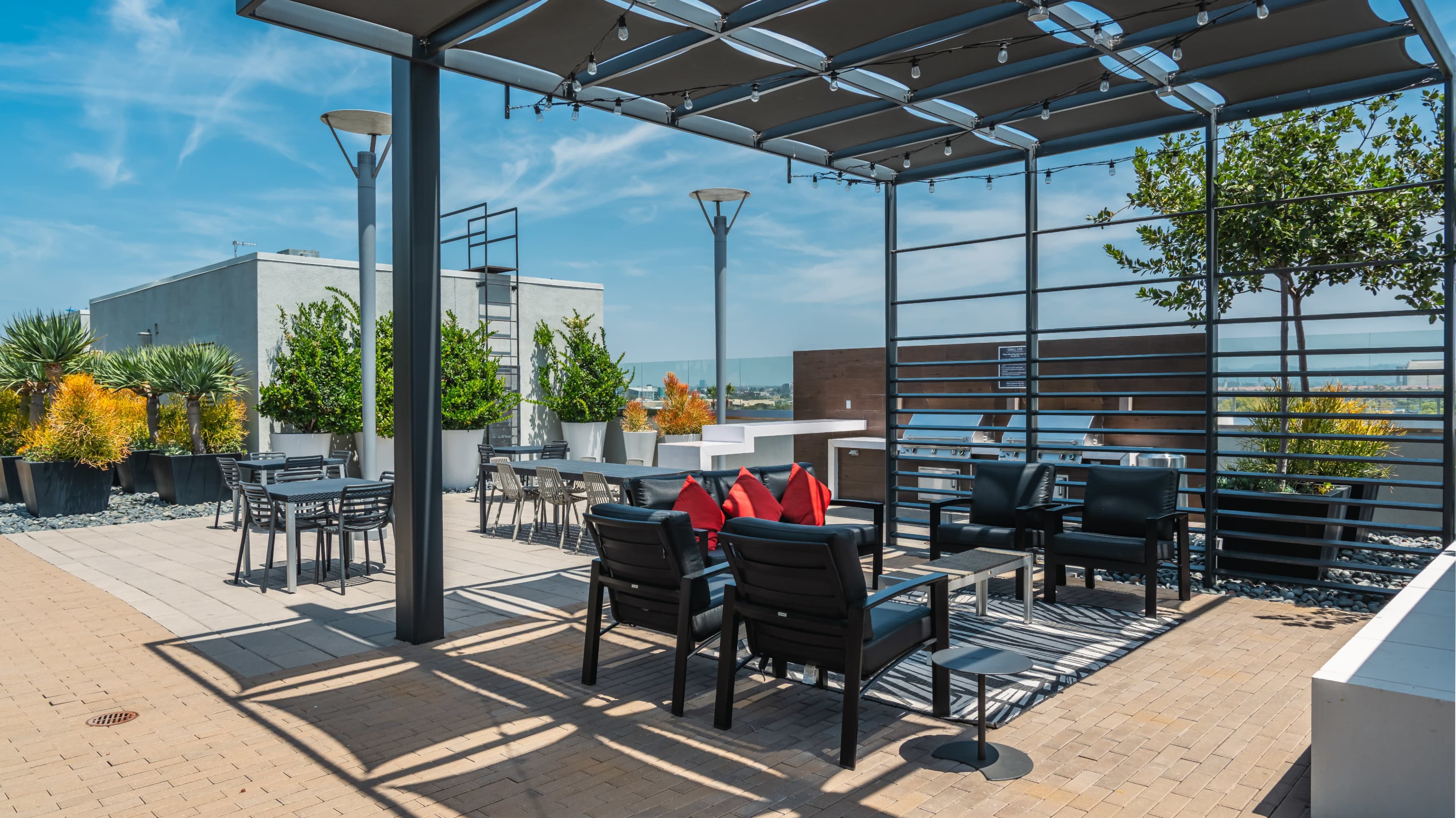 The image shows a modern rooftop terrace with seating areas, potted plants, and a pergola providing shade.