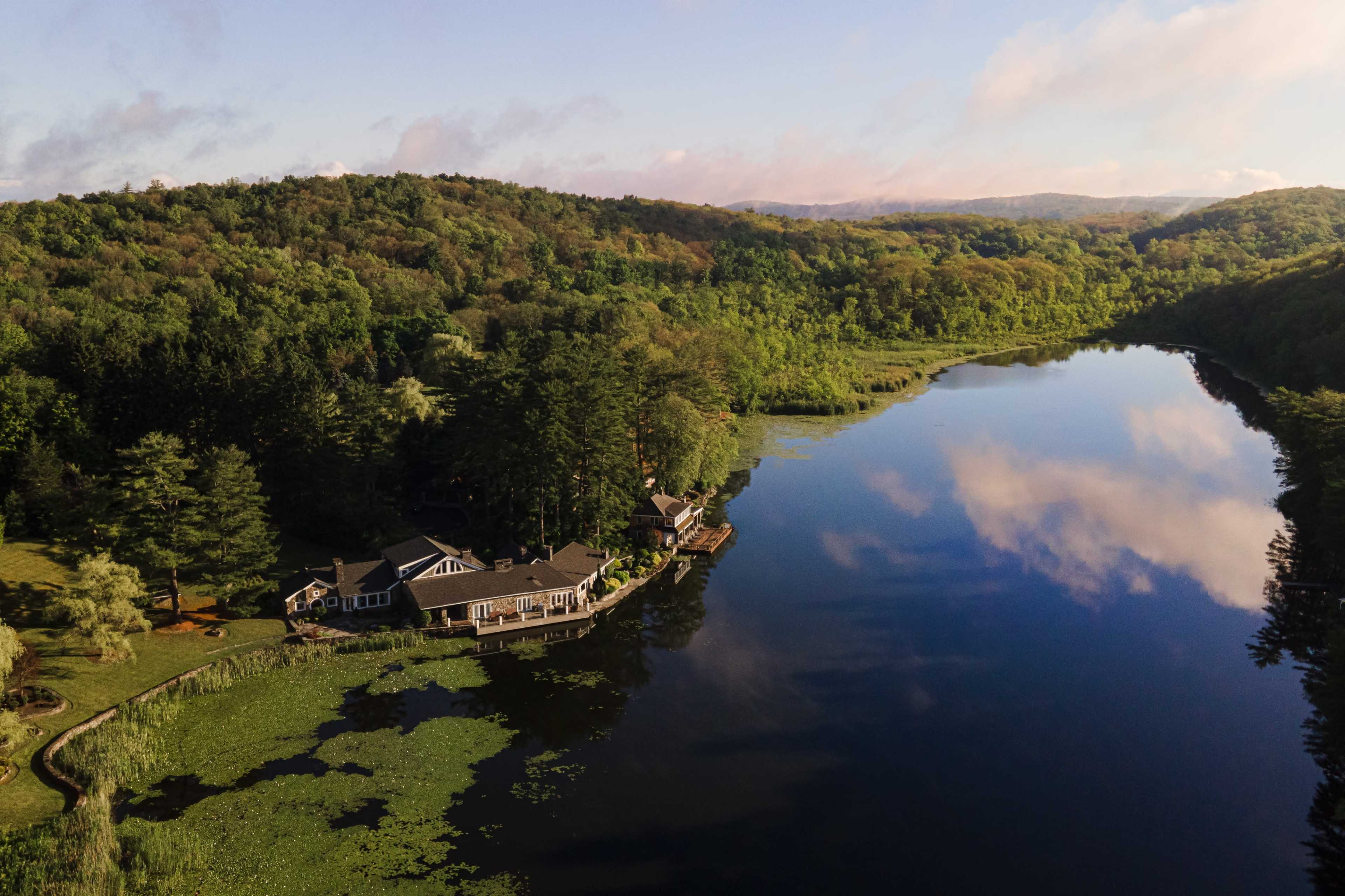 The image shows a picturesque lakeside property surrounded by lush greenery and reflections of clouds on the water's surface.