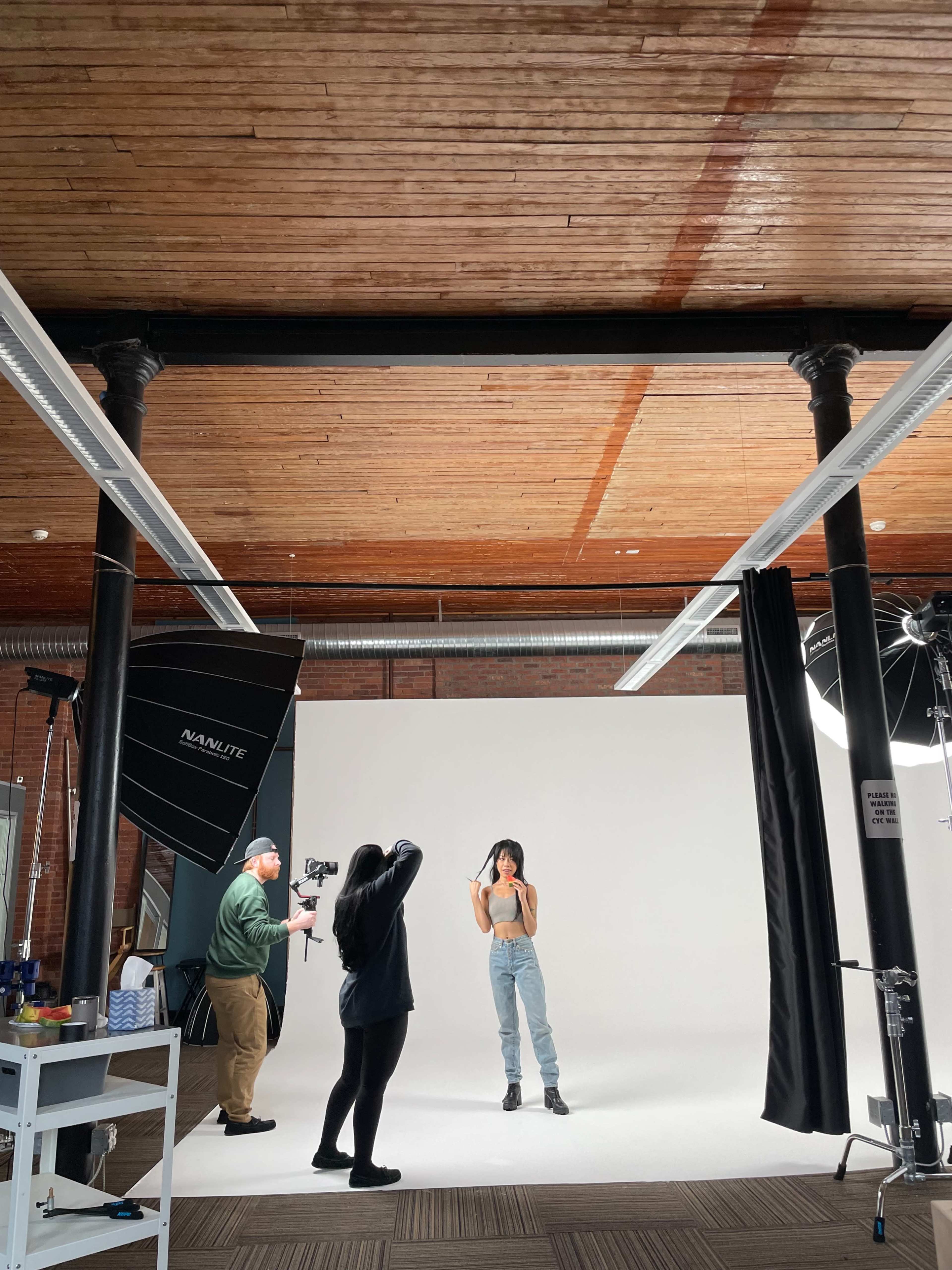A photoshoot is taking place in a studio with a model posing in front of a white backdrop while a photographer and another crew member set up equipment.