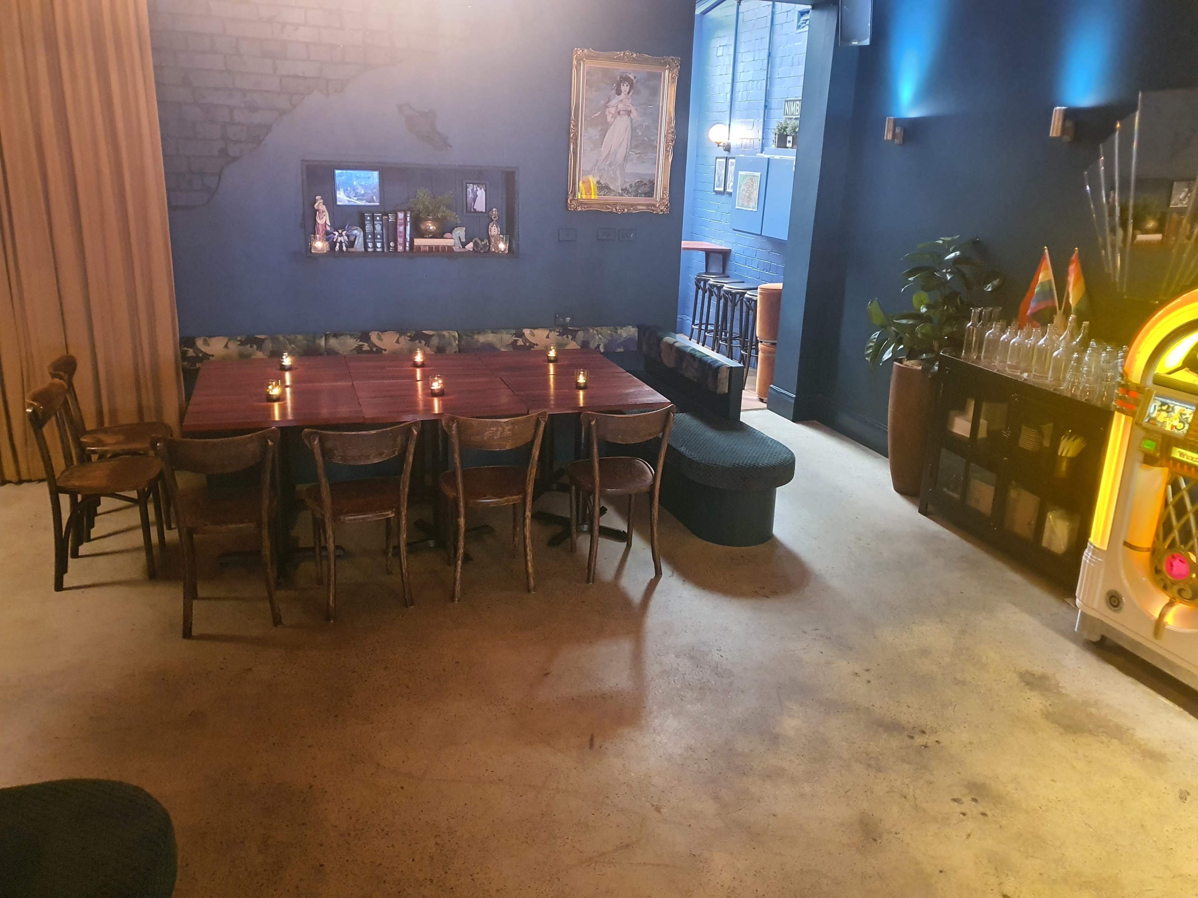 A dimly lit room with a long wooden table surrounded by chairs, a corner seating area, and a jukebox against a blue wall.