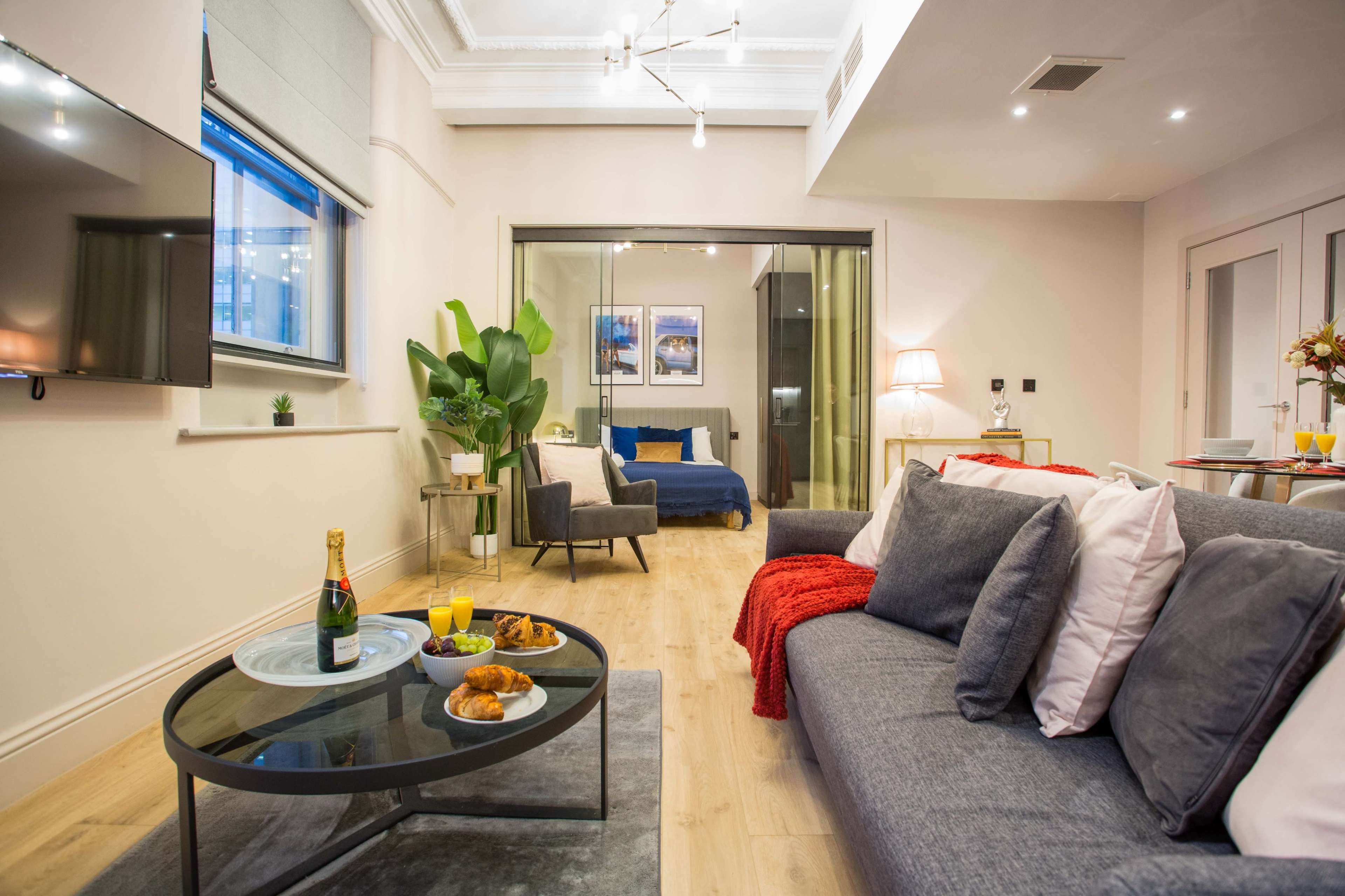 The image shows a modern living room with a grey sofa facing a glass coffee table, featuring a bottle of champagne and snacks, and a view into a bedroom area with a blue bed and framed pictures on the wall.