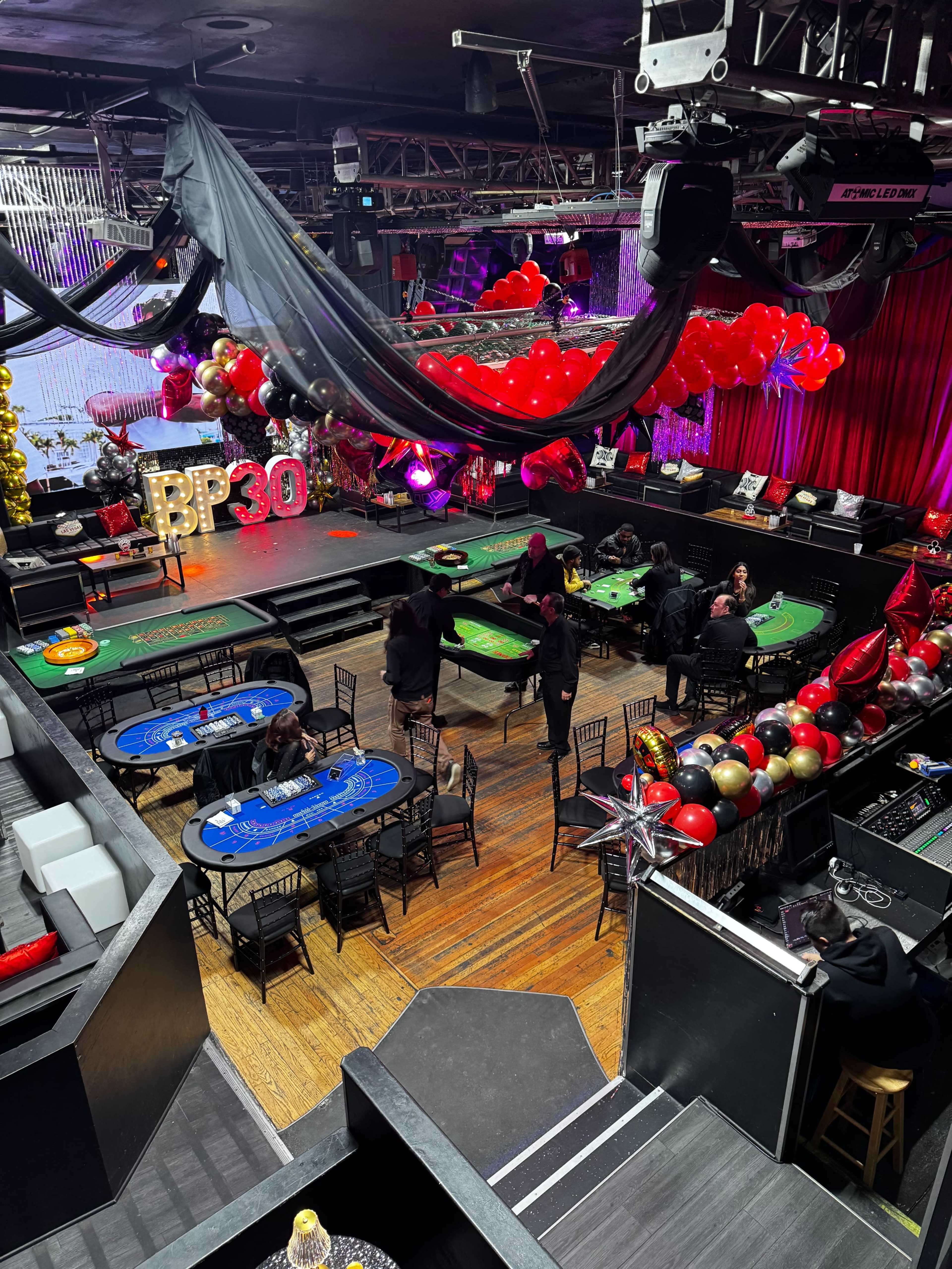The image shows a decorated venue with gaming tables and colorful balloons for a celebration event.
