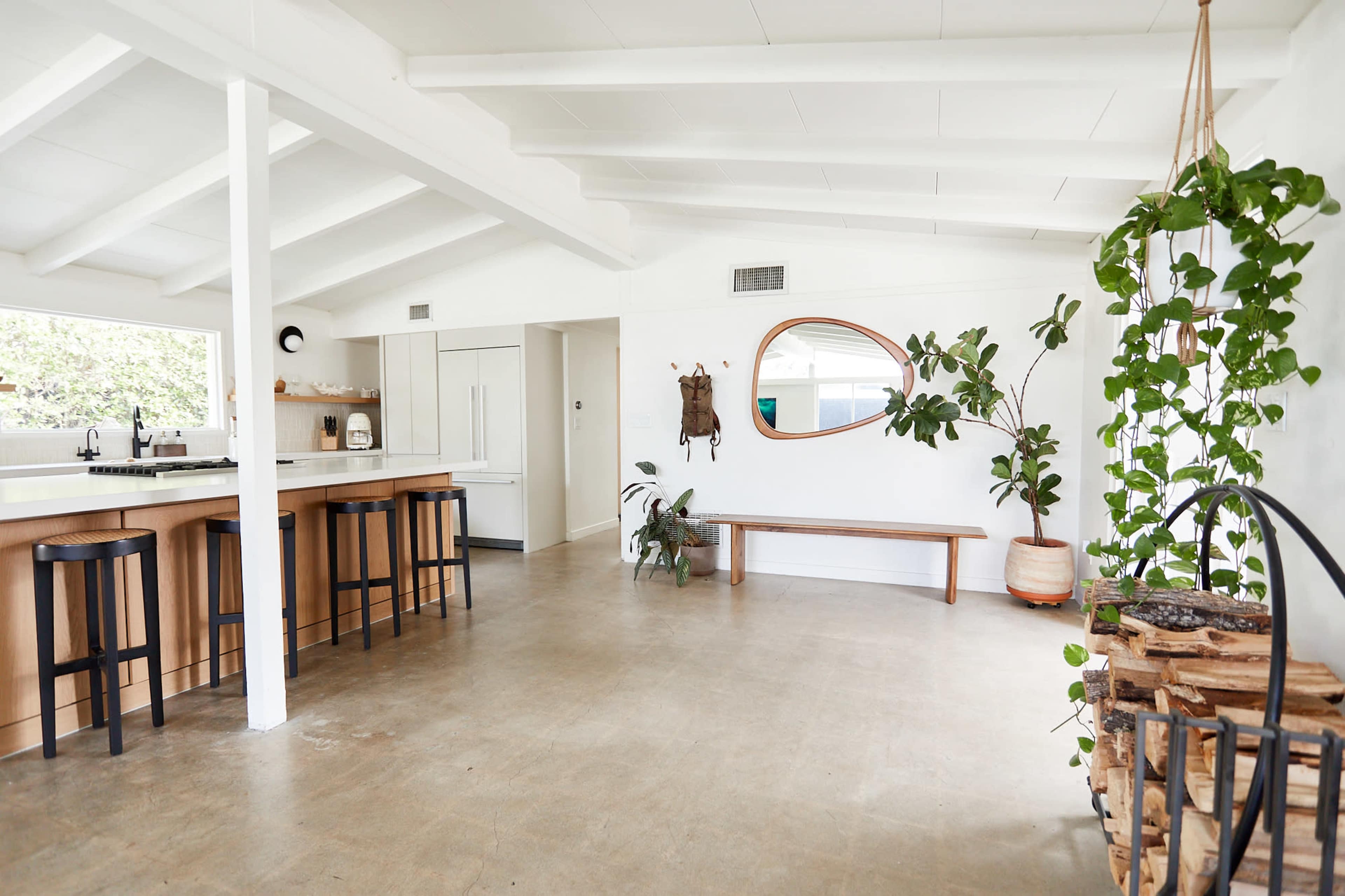 The image shows a spacious interior room with a kitchen area featuring a long countertop, a round mirror on the wall, a bench, and various plants.