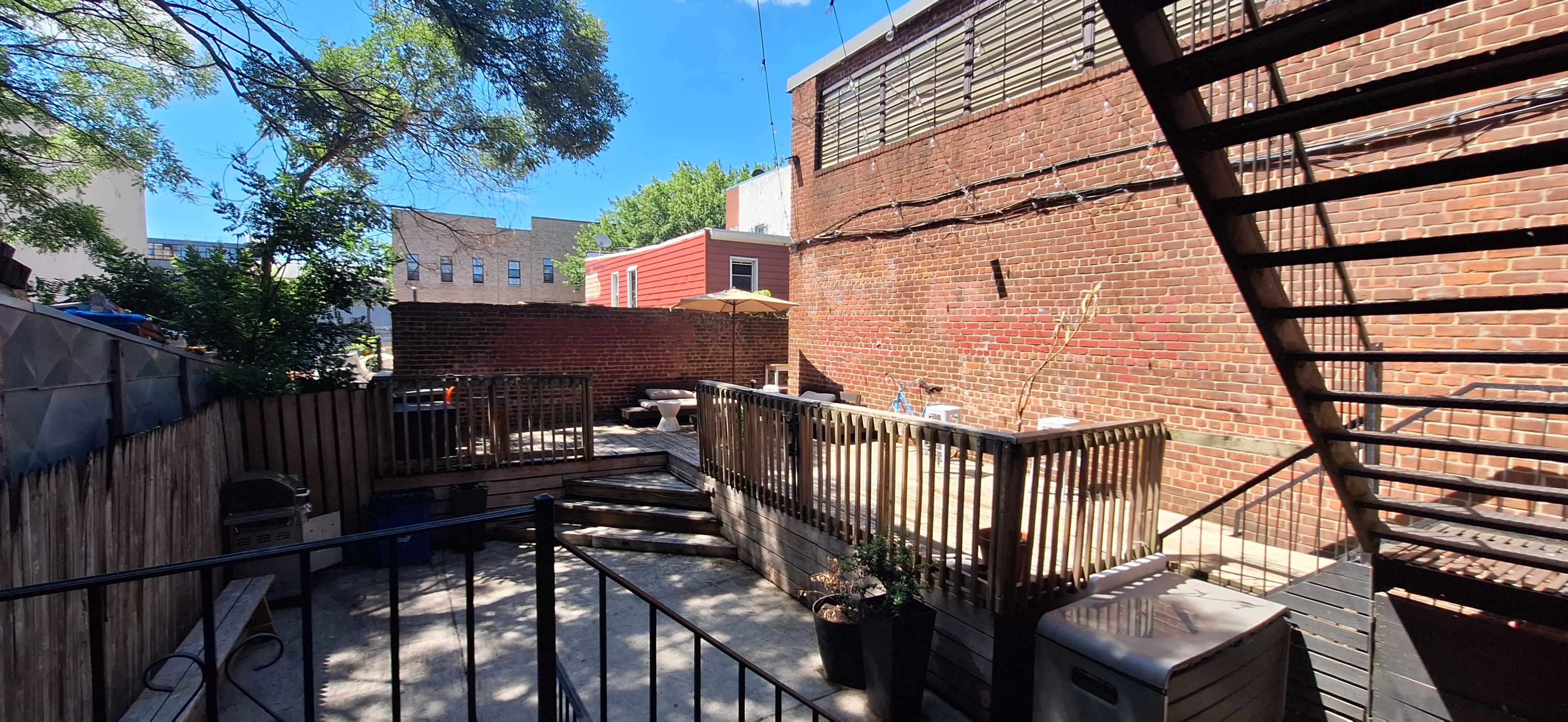 The image shows a sunlit backyard area featuring wooden decks, brick walls, and an outdoor umbrella.