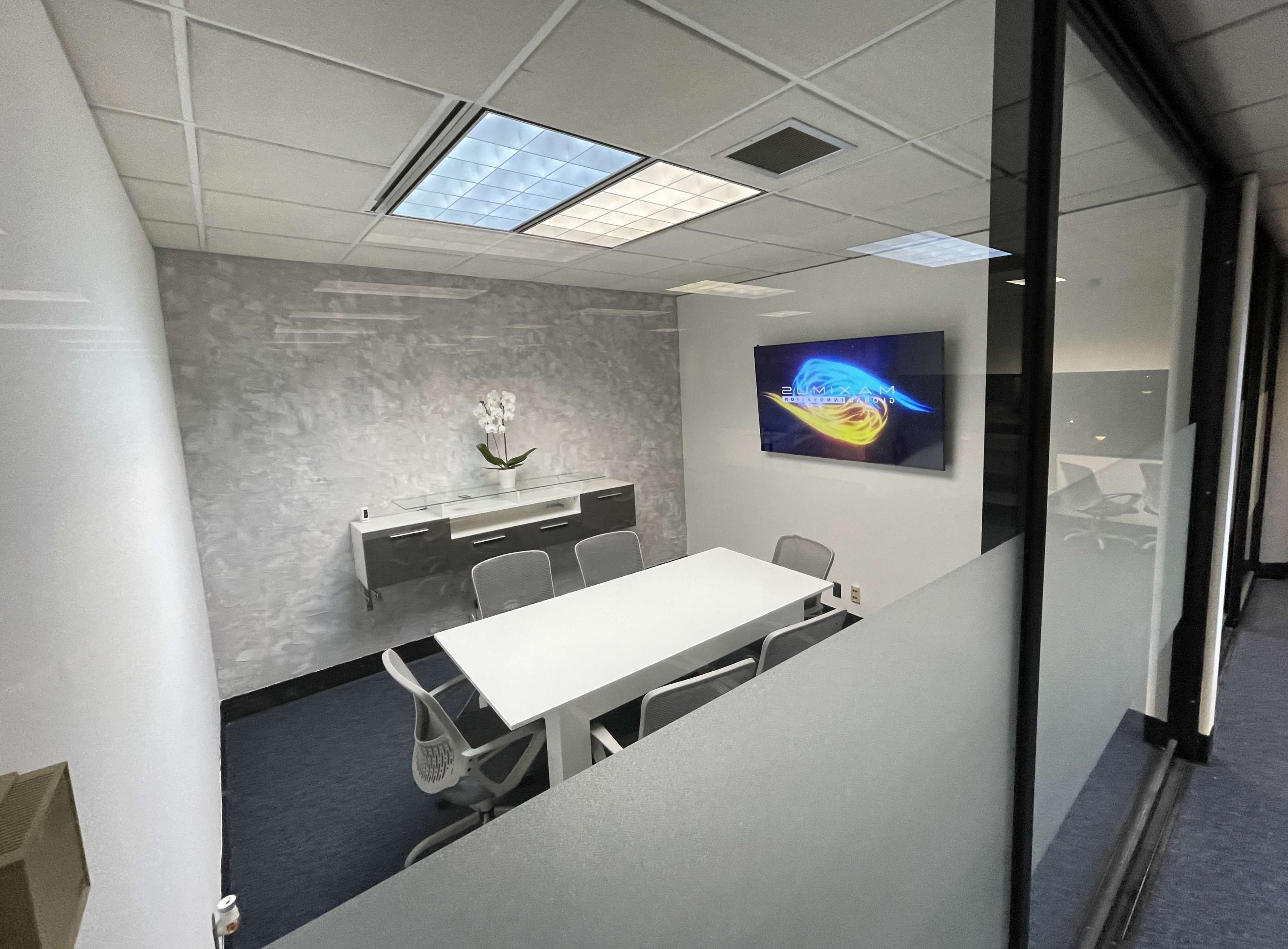 A modern conference room with a white table, six chairs, a wall-mounted television, and a decorative plant in a glass-walled space.