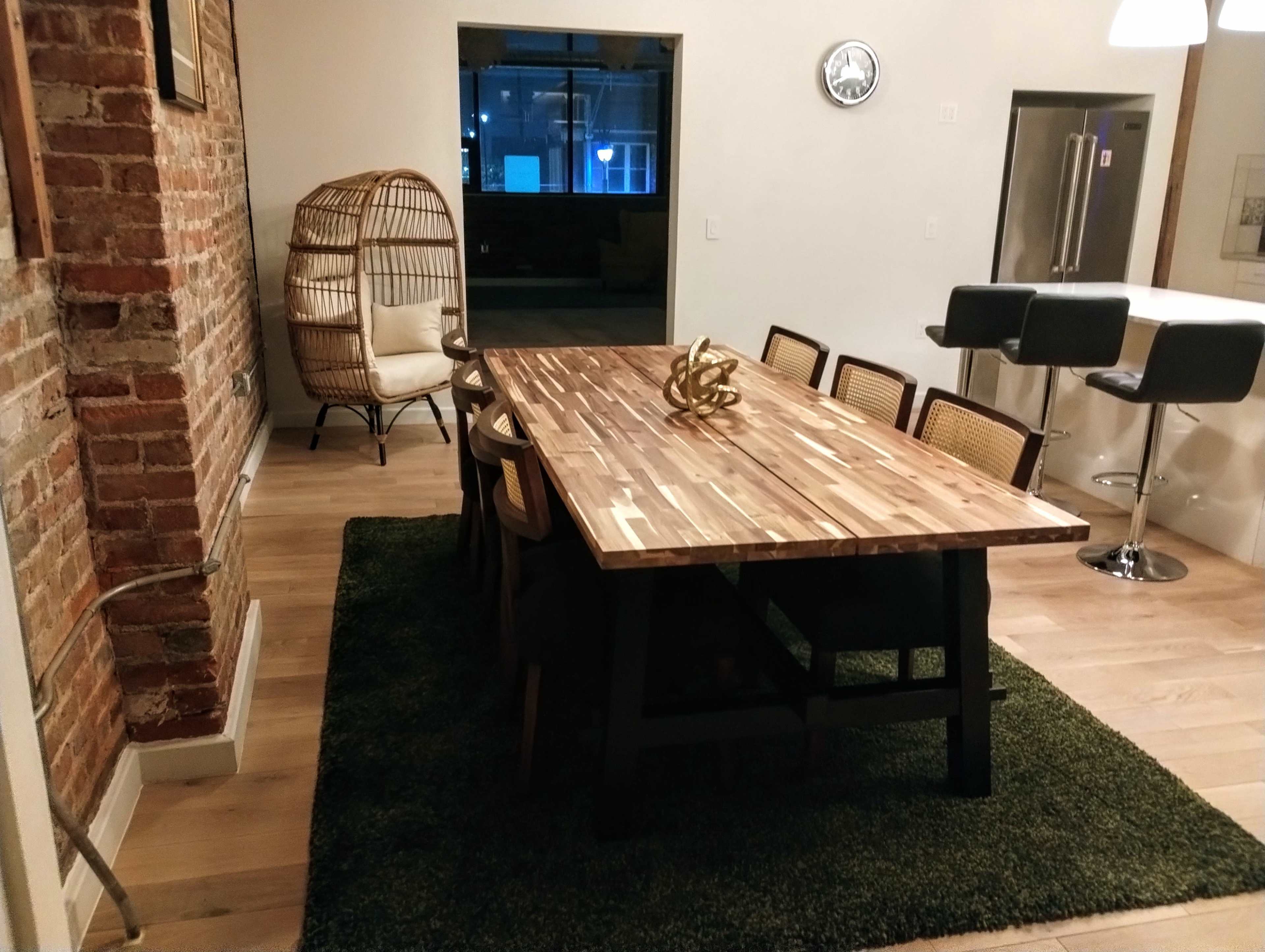 A modern dining area featuring a wooden table surrounded by eight chairs, a woven chair in the corner, and a rug on the floor, with exposed brick walls and a kitchen area in the background.