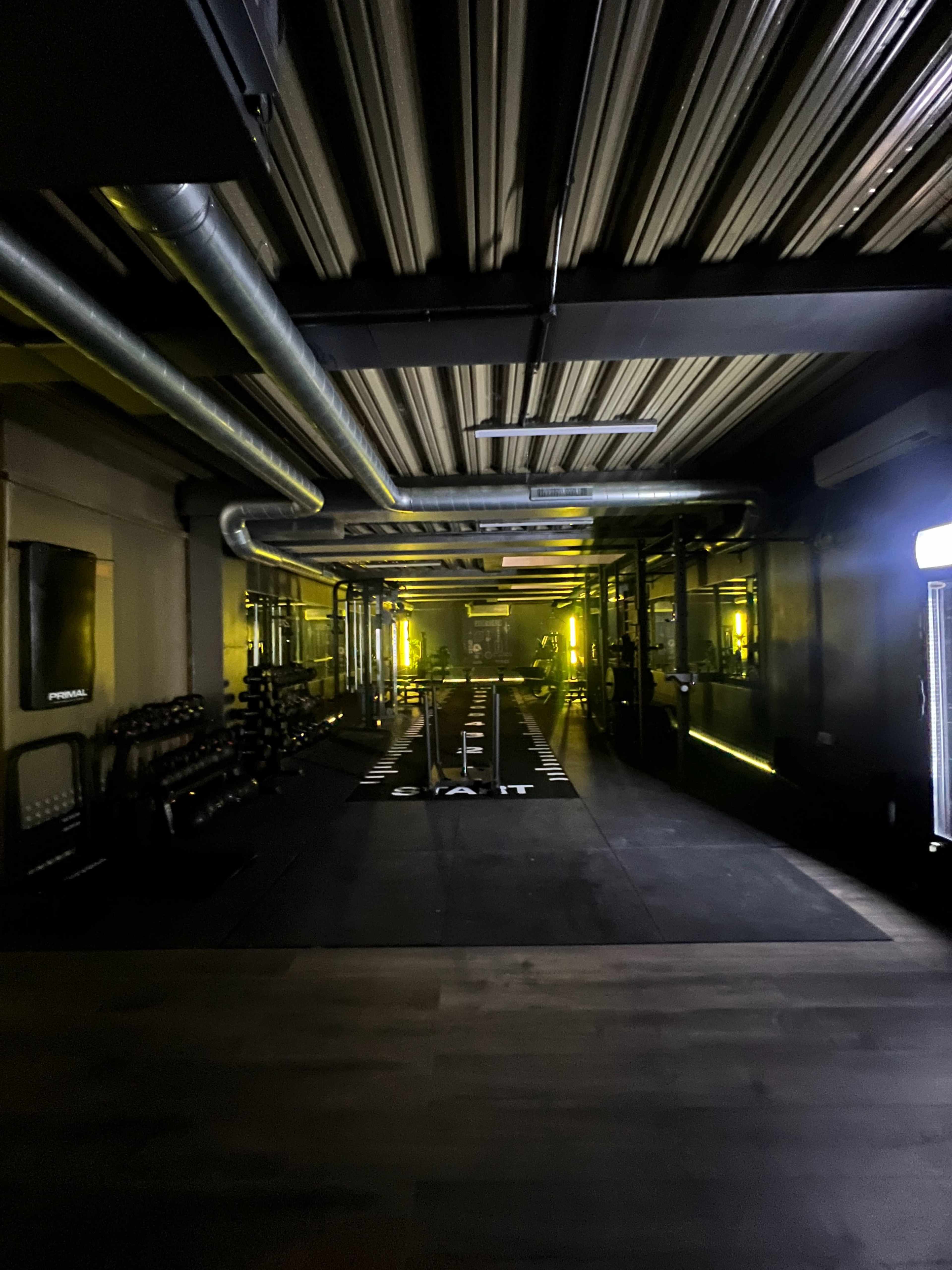 A dimly lit gym with equipment lined up along the walls and a central training area marked by floor mats.
