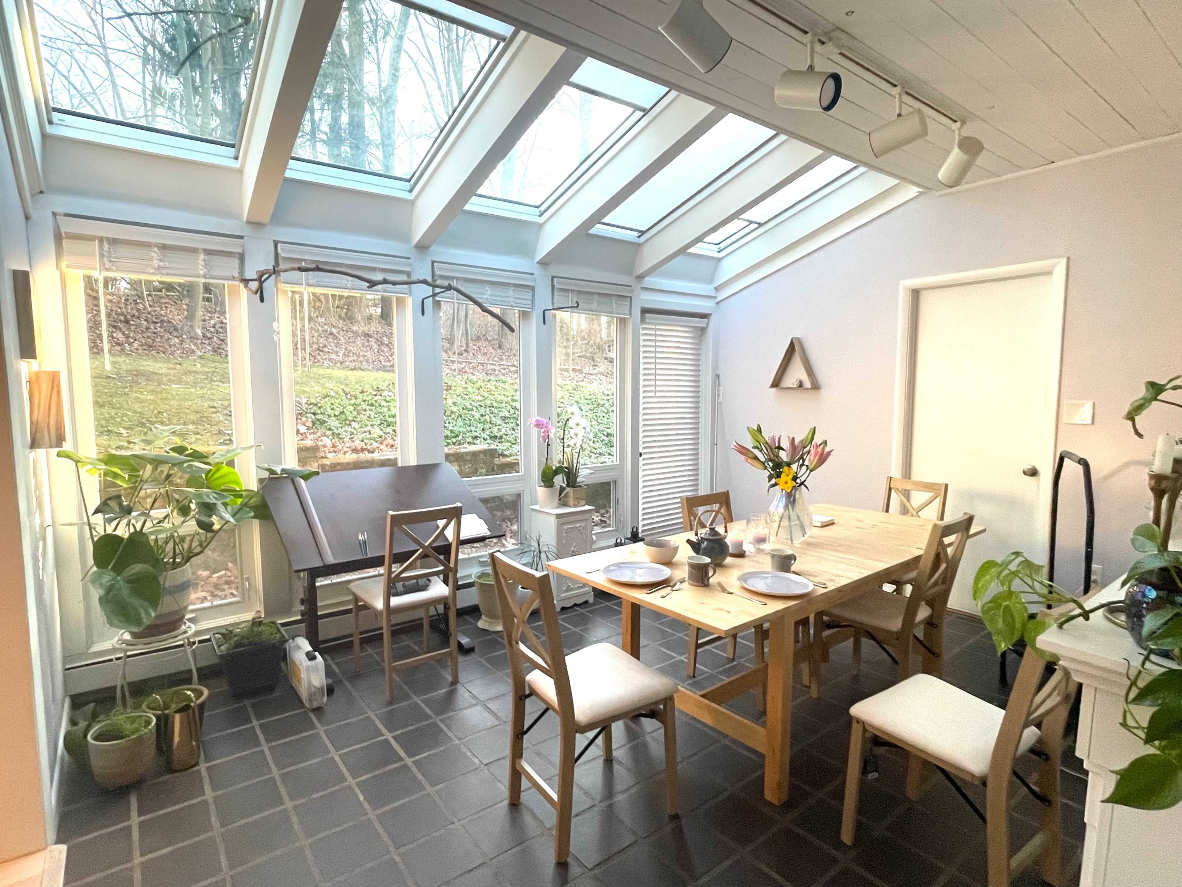 A bright dining area with large skylights, a wooden table set for a meal, and plants in the corners.
