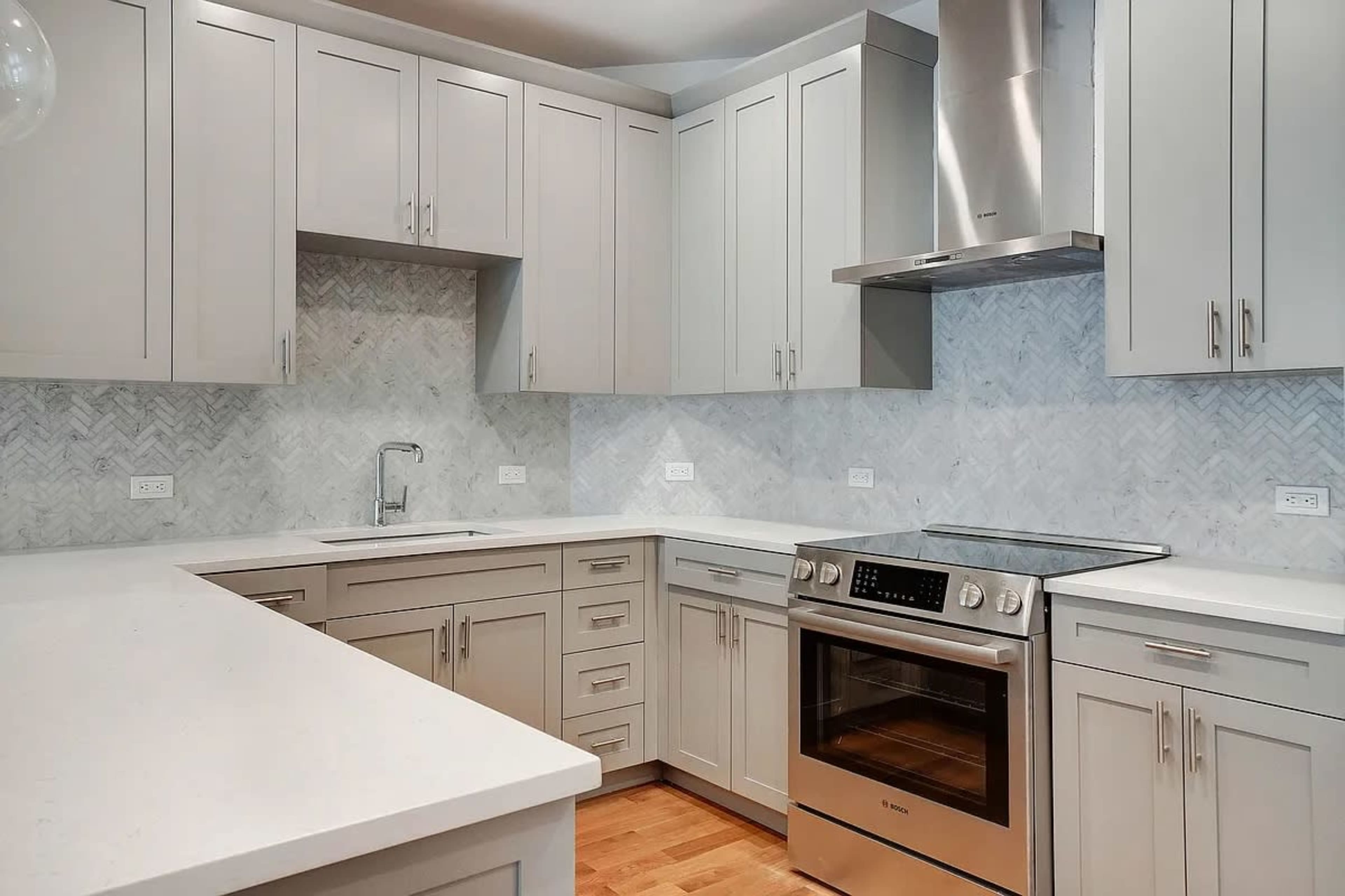 A modern kitchen features light gray cabinetry, a tile backsplash in a herringbone pattern, and stainless steel appliances.