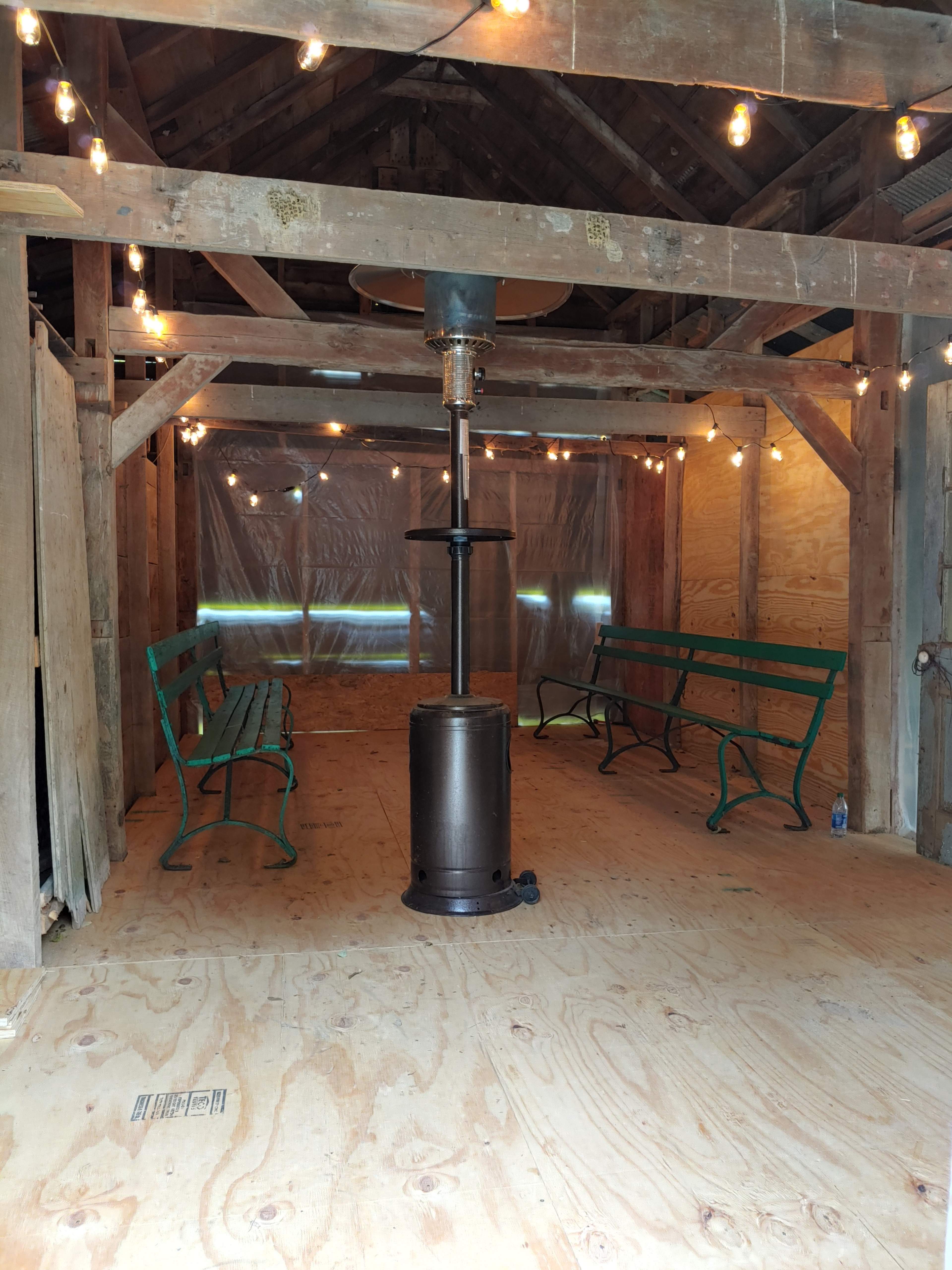 The image shows an interior space with wooden beams and flooring, featuring a central heater and green benches along the sides, illuminated by string lights.