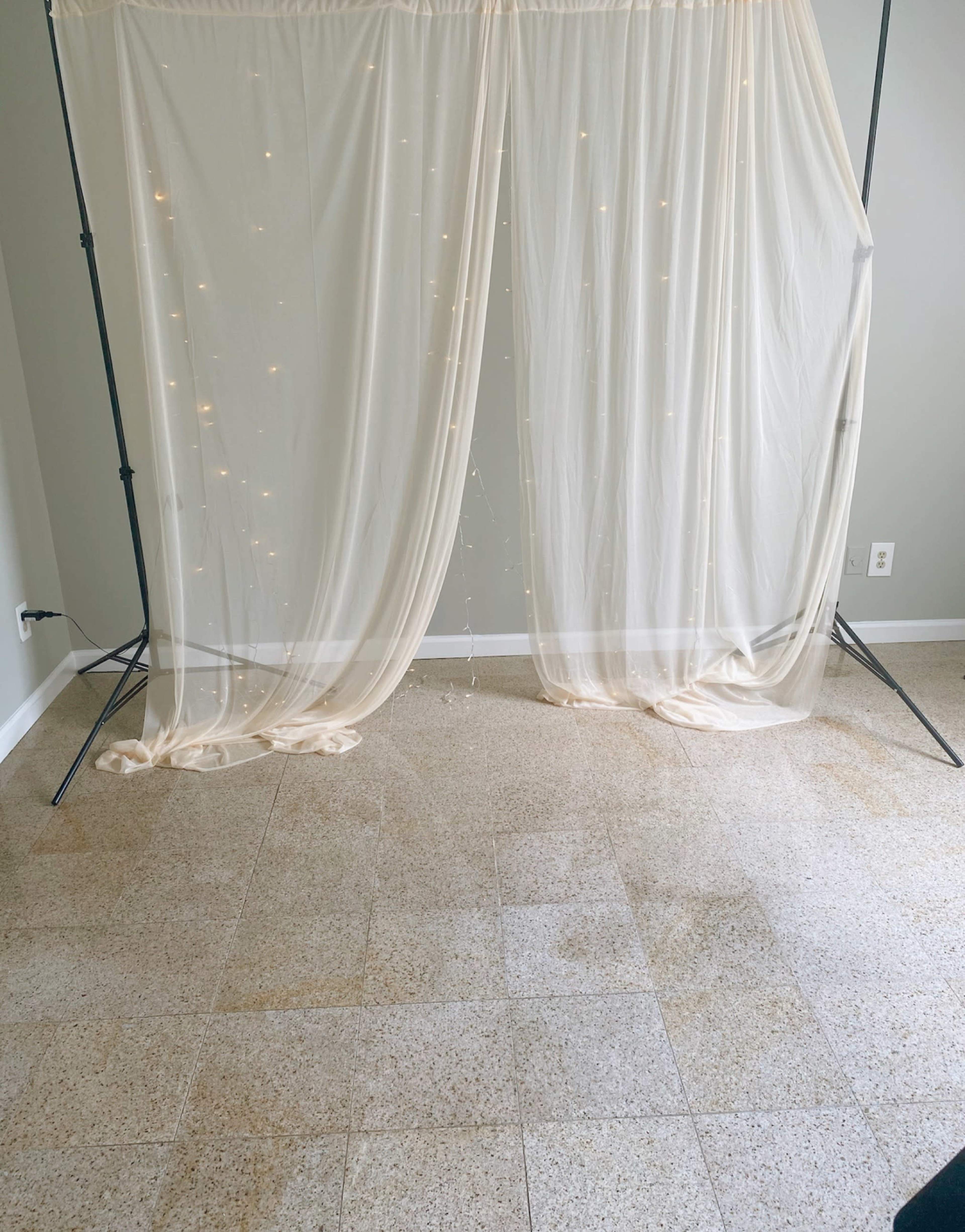 A lightly draped sheer curtain with string lights is set up on a frame against a plain wall in an empty room.