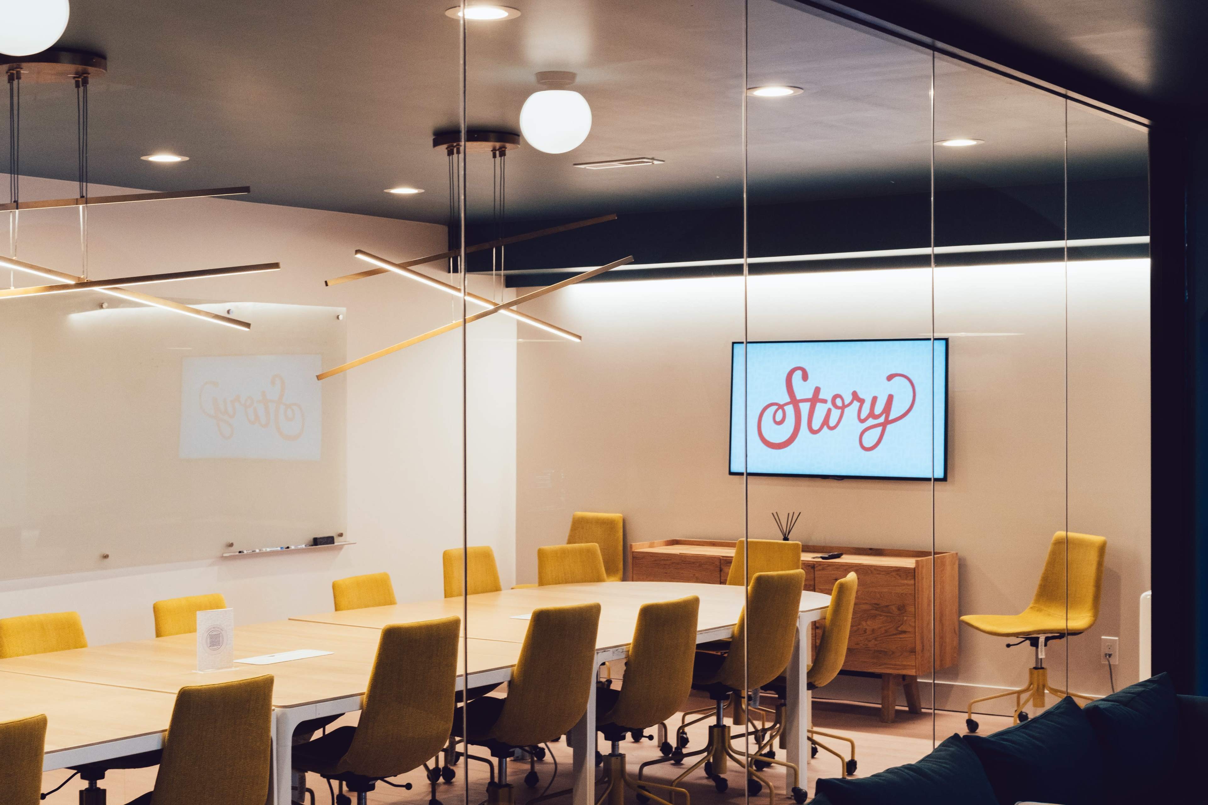 A modern conference room features a large table surrounded by yellow chairs, with a screen displaying the word "Story" on the wall.