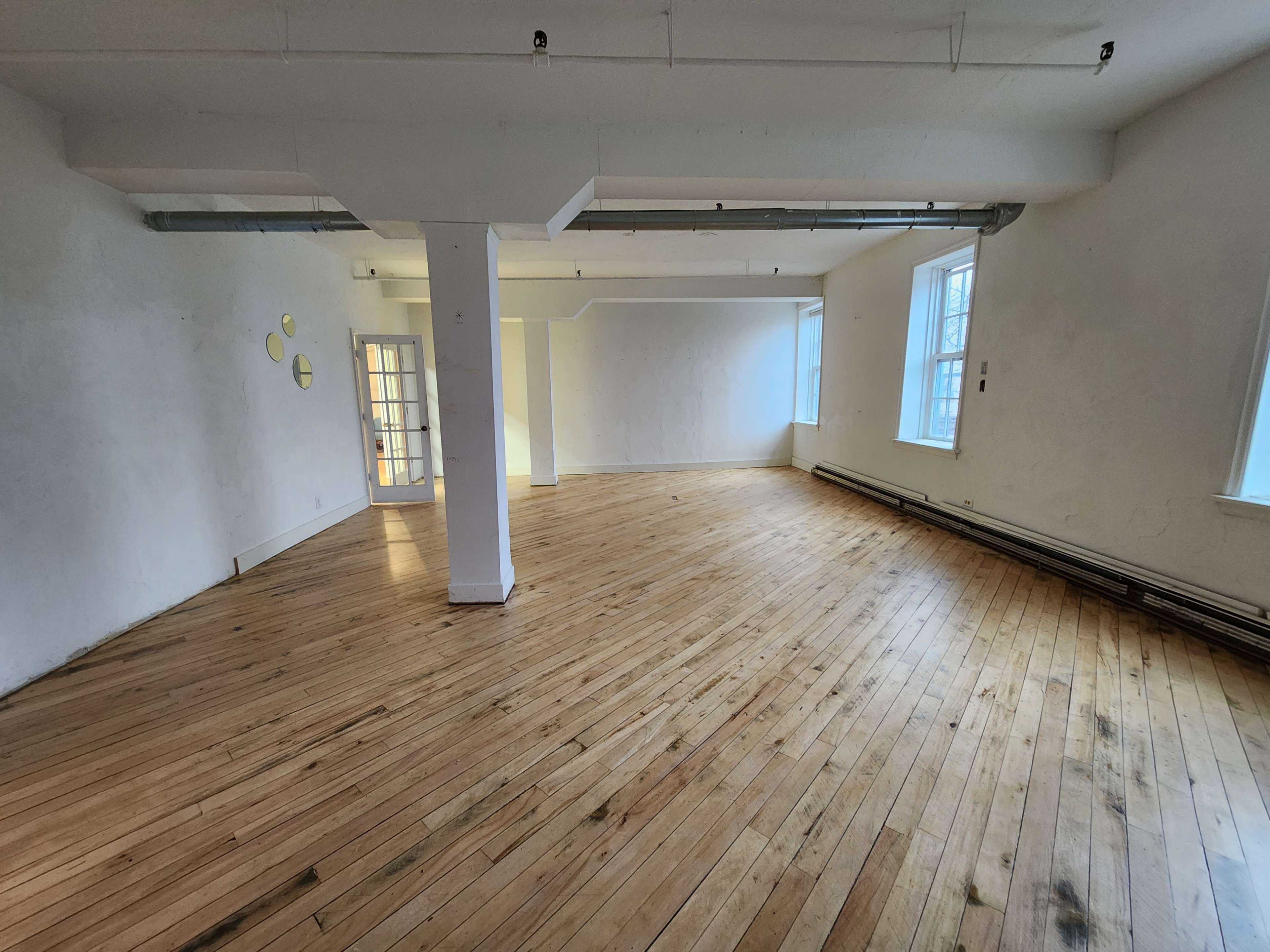 A spacious, empty room with wooden flooring, two windows, and a column in the center.