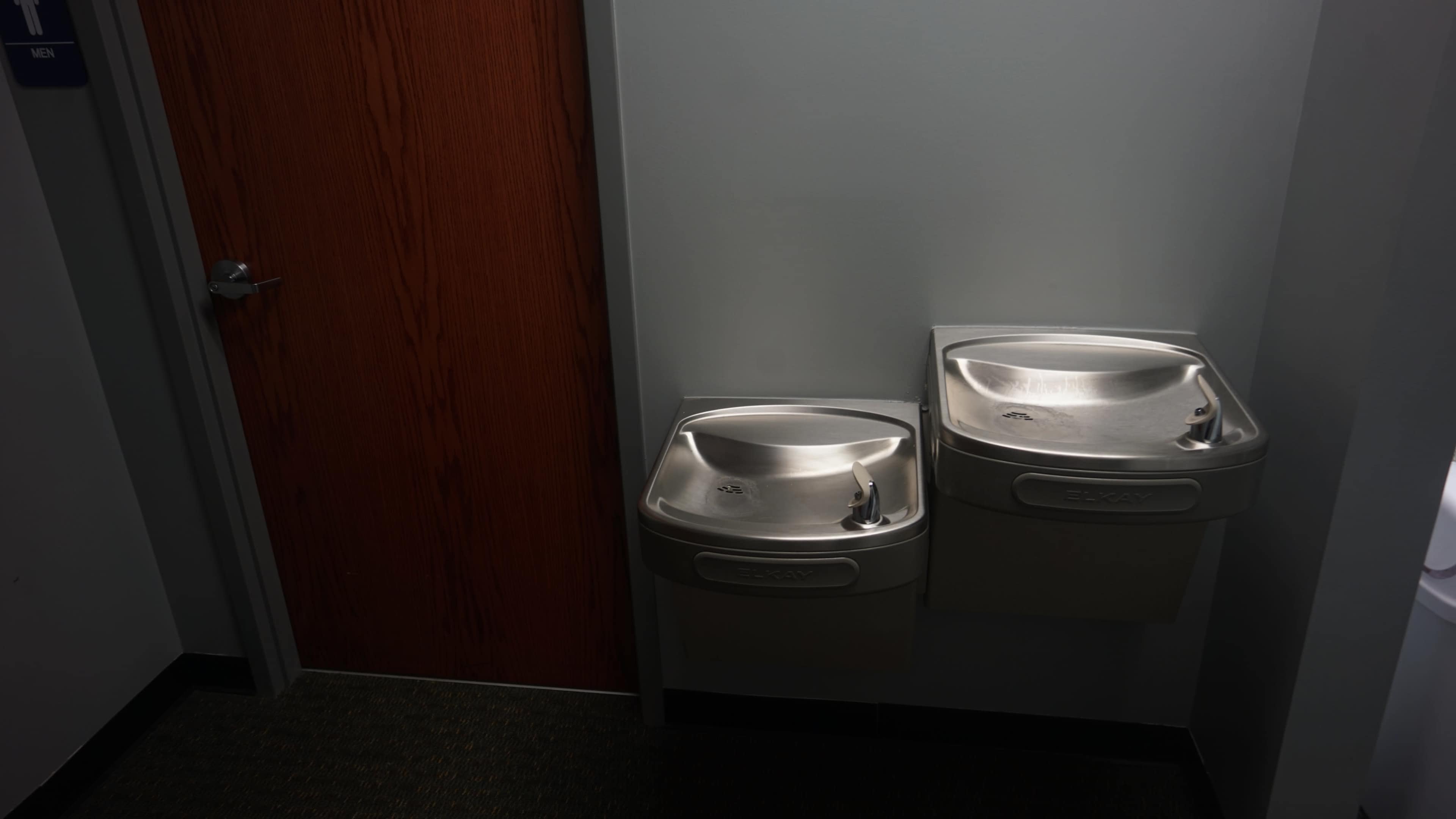 Two stainless steel drinking fountains mounted on a wall next to a wooden door labeled "Men."