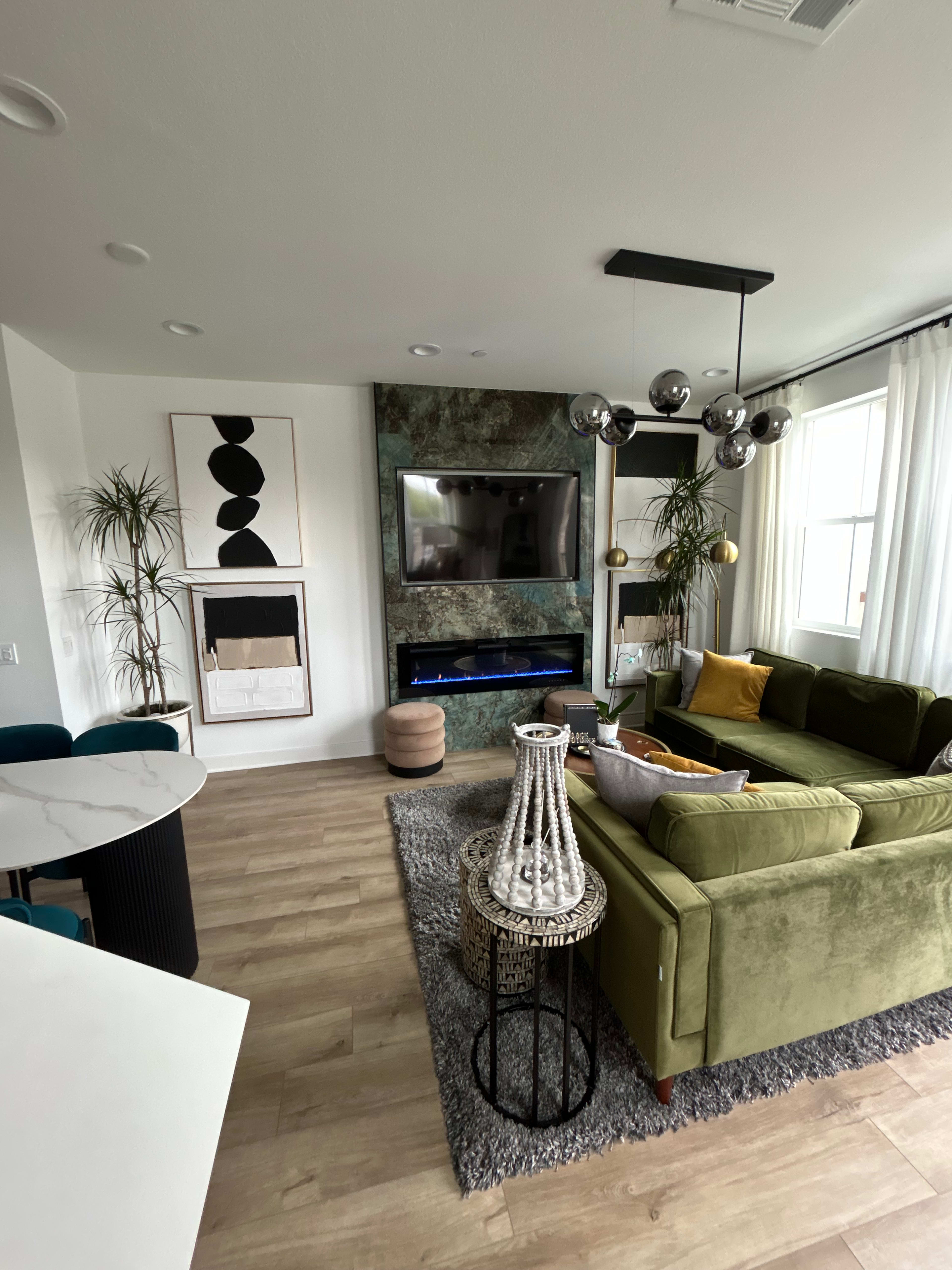 The image shows a modern living room with a green sofa, a round coffee table, a large wall-mounted TV, and various decorative plants.